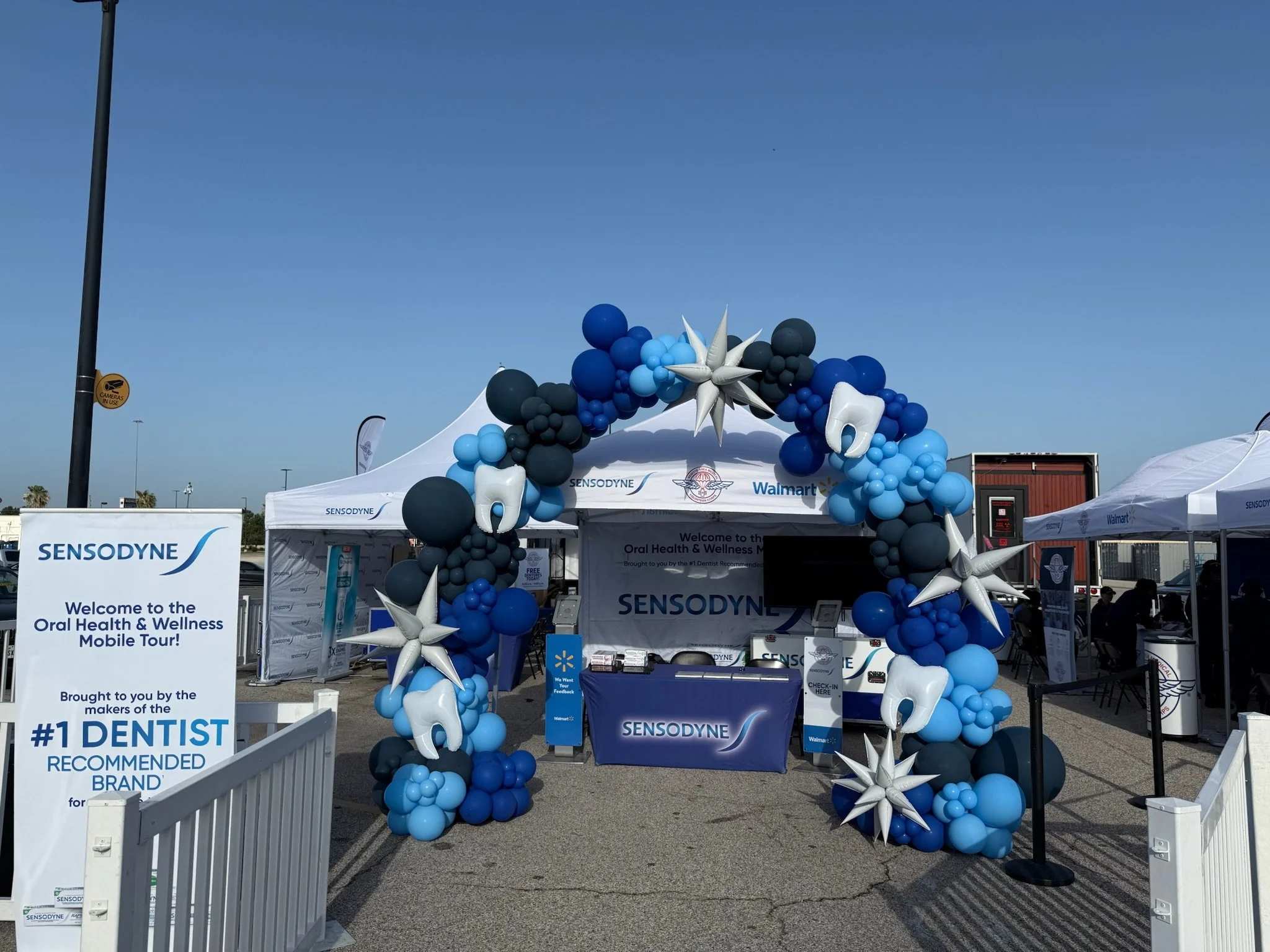 Sensodyne booth at an event decorated with blue, black, and white balloons, including balloon teeth and star-shaped balloons, set up outdoors on a sunny day with clear sky.