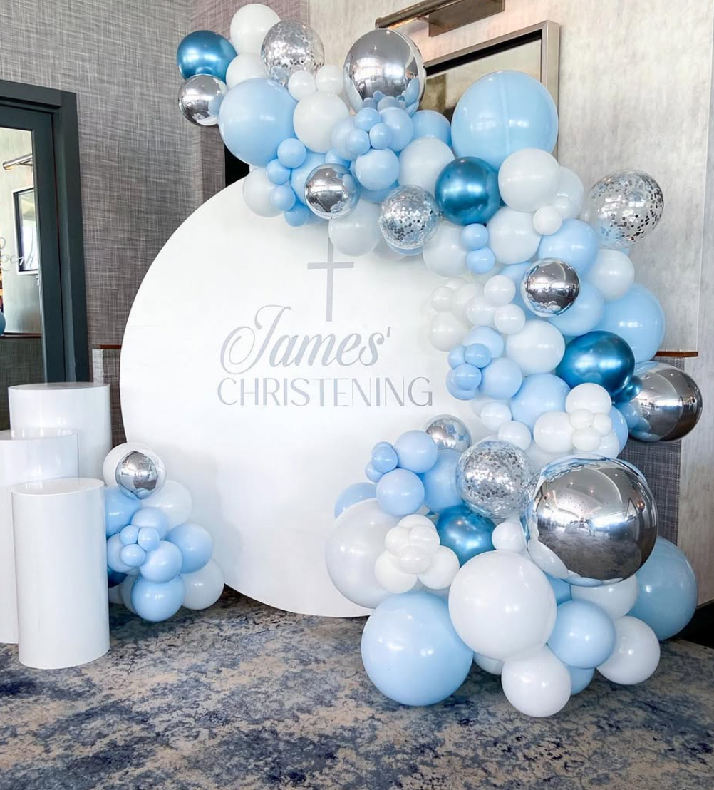 Balloon arrangement with blue, white, silver, and confetti balloons surrounding a white circular backdrop for a christening celebration for James.