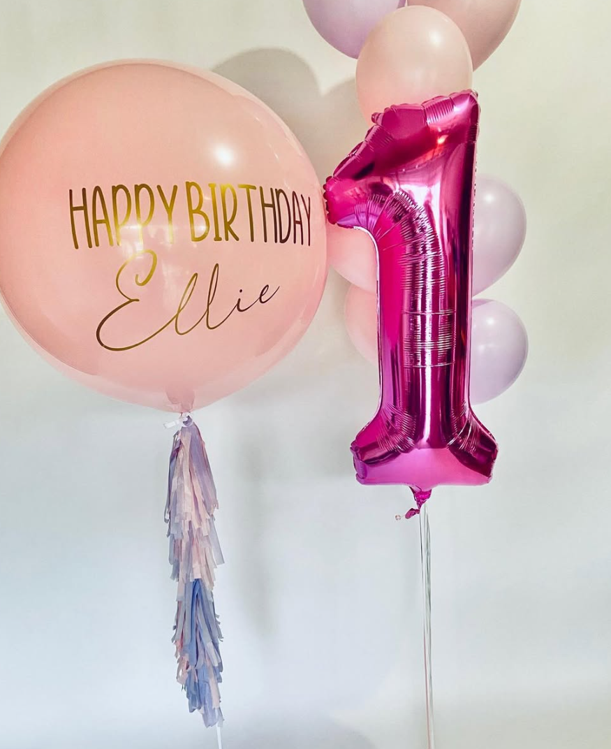 Pink birthday balloon with the message 'Happy Birthday Ellie' in gold and black lettering, and pink balloons including a shiny pink number one balloon, set against a light background.