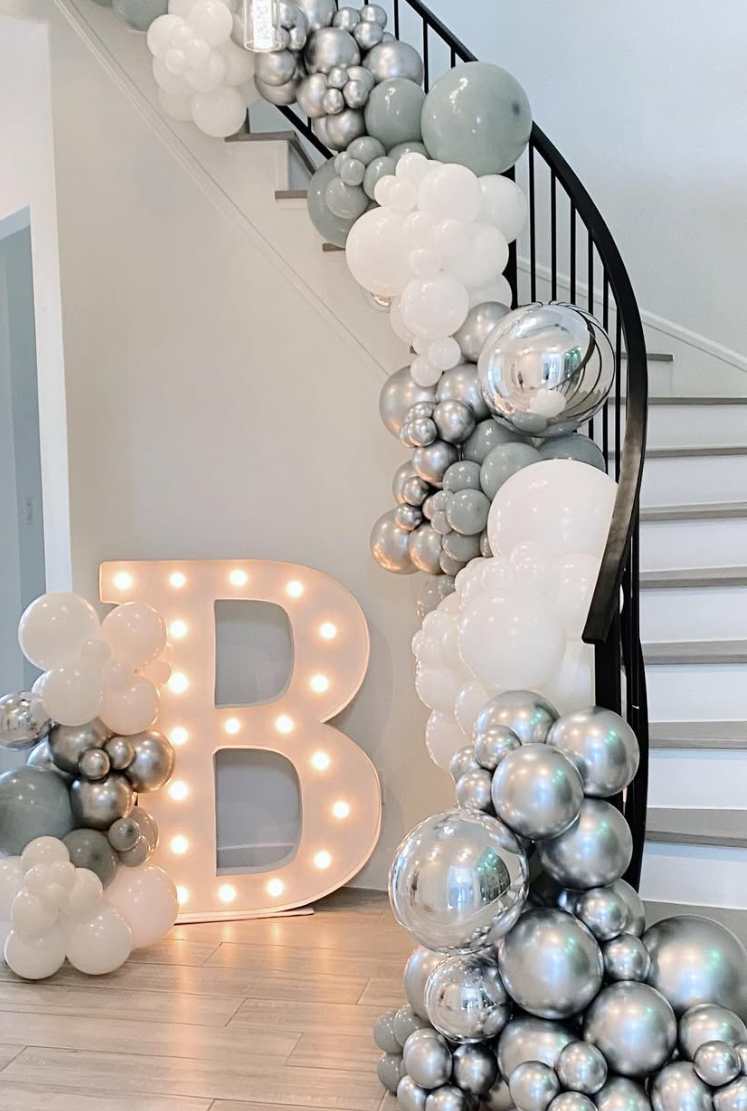 Decorative balloon arrangement with silver, white, and gray balloons along a staircase, and a large illuminated letter 'B' with light bulbs near the bottom of the staircase.