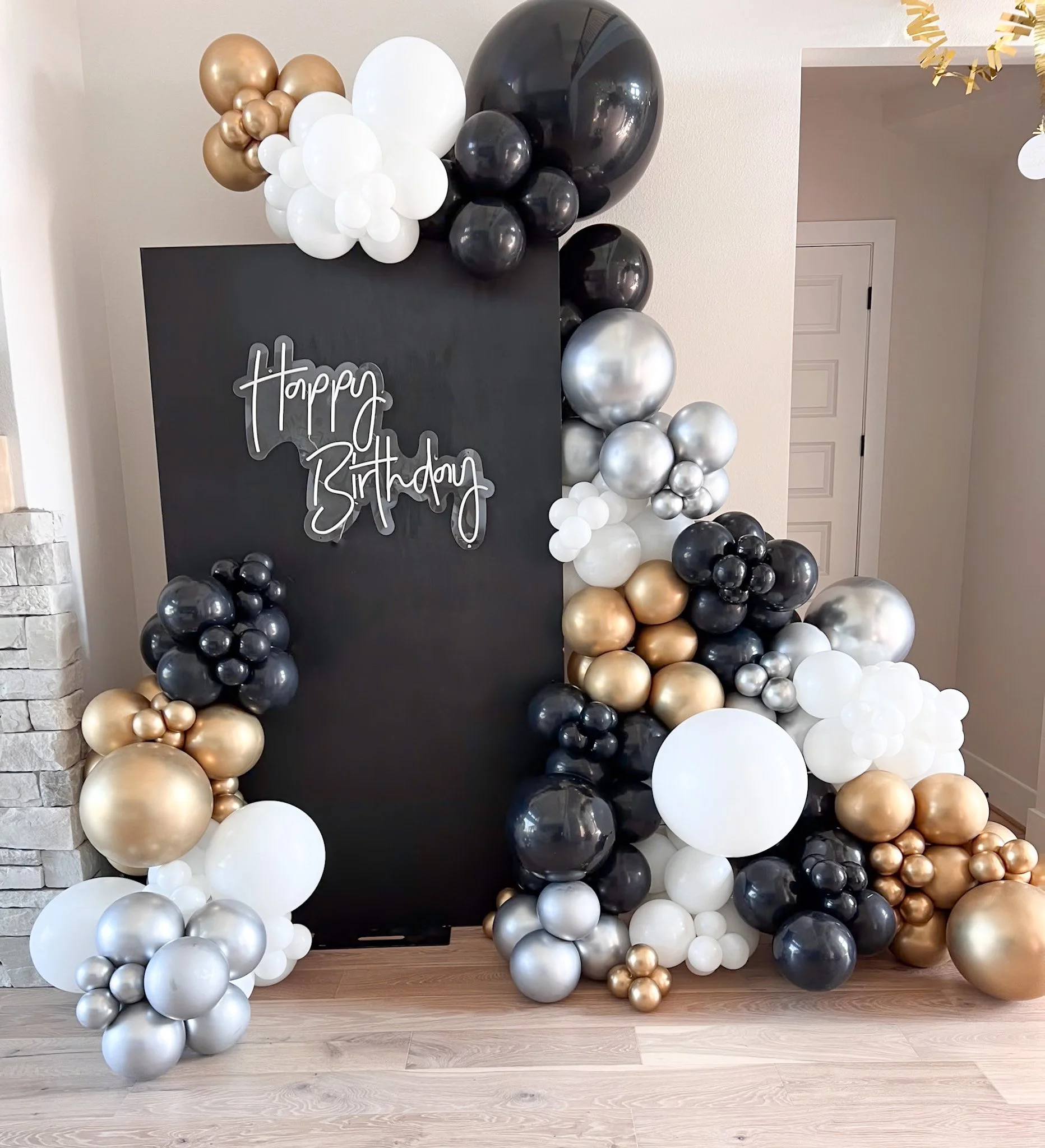 Blackboard with a balloon arch and cluster decorations in gold, silver, black, and white for a birthday celebration.