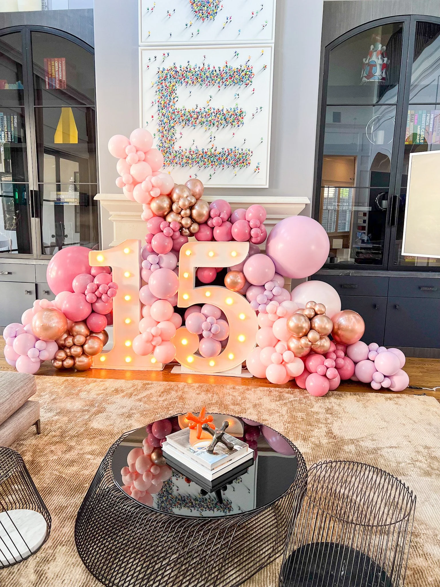 Decorative birthday display with a large illuminated number 15, pink, white, and metallic balloons, and a framed letter E made of small colorful pins on the wall.