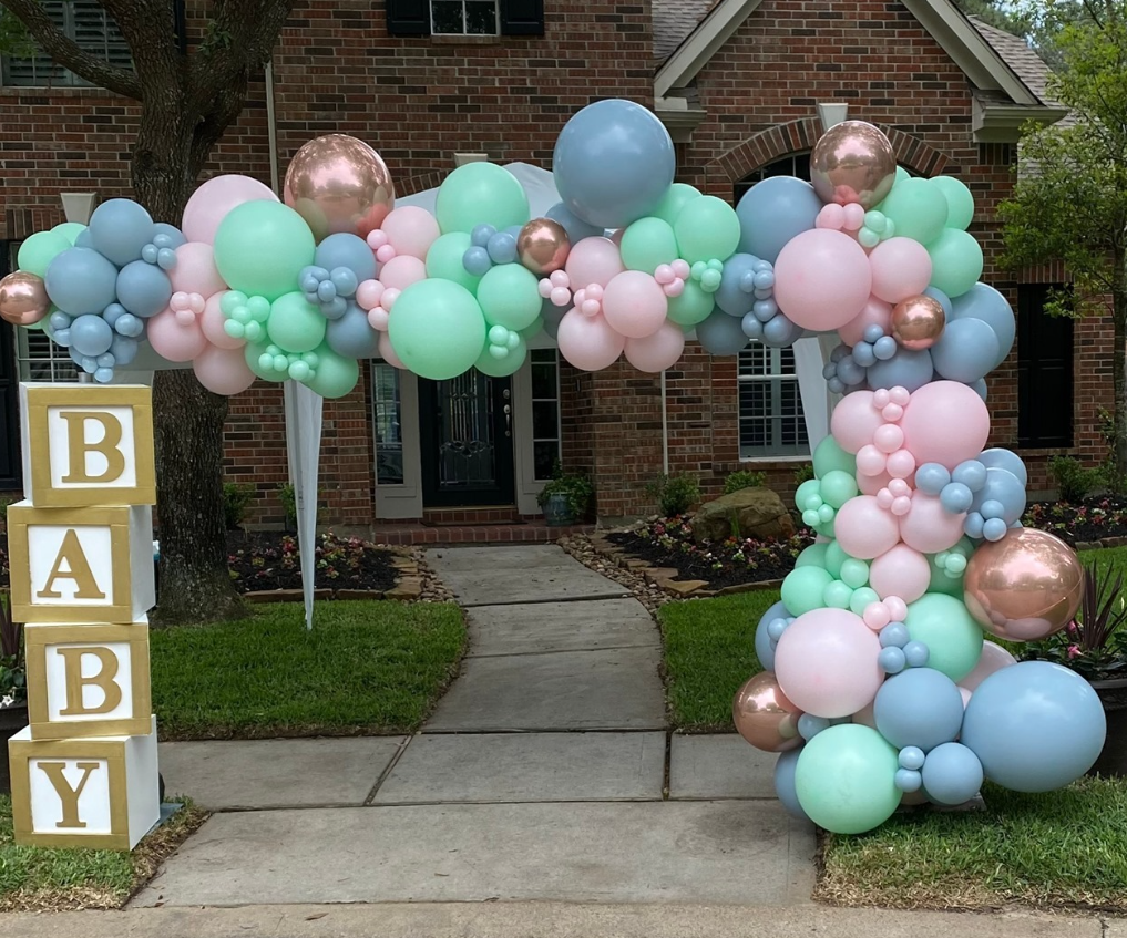 Balloon arch decorated with pastel pink, blue, green, and metallic rose gold balloons in front of a brick house with a sidewalk leading to the door. To the left, a stack of four gold-colored blocks spells out 'BABY' in white letters.