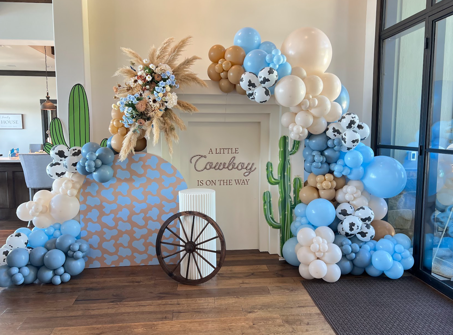 Decorative arch with blue, cream, and black cow-print balloons, and cactus cutouts. A floral arrangement with pampas grass and flowers on top. A sign in the middle reads "A LITTLE Cowboy IS ON THE WAY." Wooden wagon wheel and white column in front of