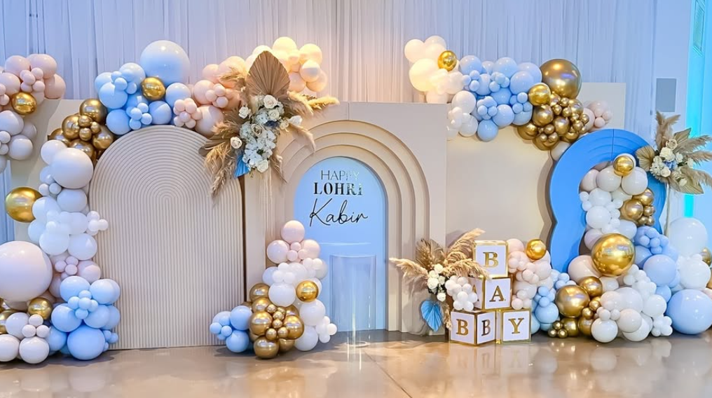 Decorative backdrop for a baby shower with balloons in pastel pink, blue, white, and gold, floral arrangements, and blocks spelling out "BABY."
