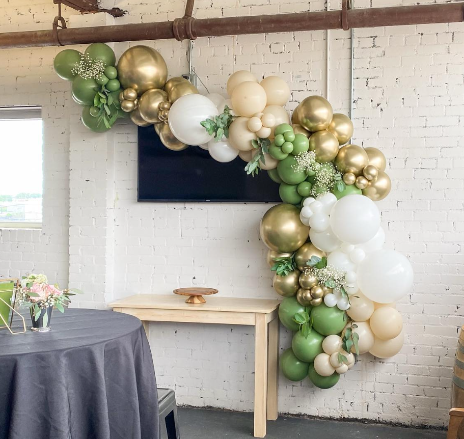 A decorative balloon arch in green, white, cream, and gold colors with greenery, set against a white brick wall in an indoor event space.