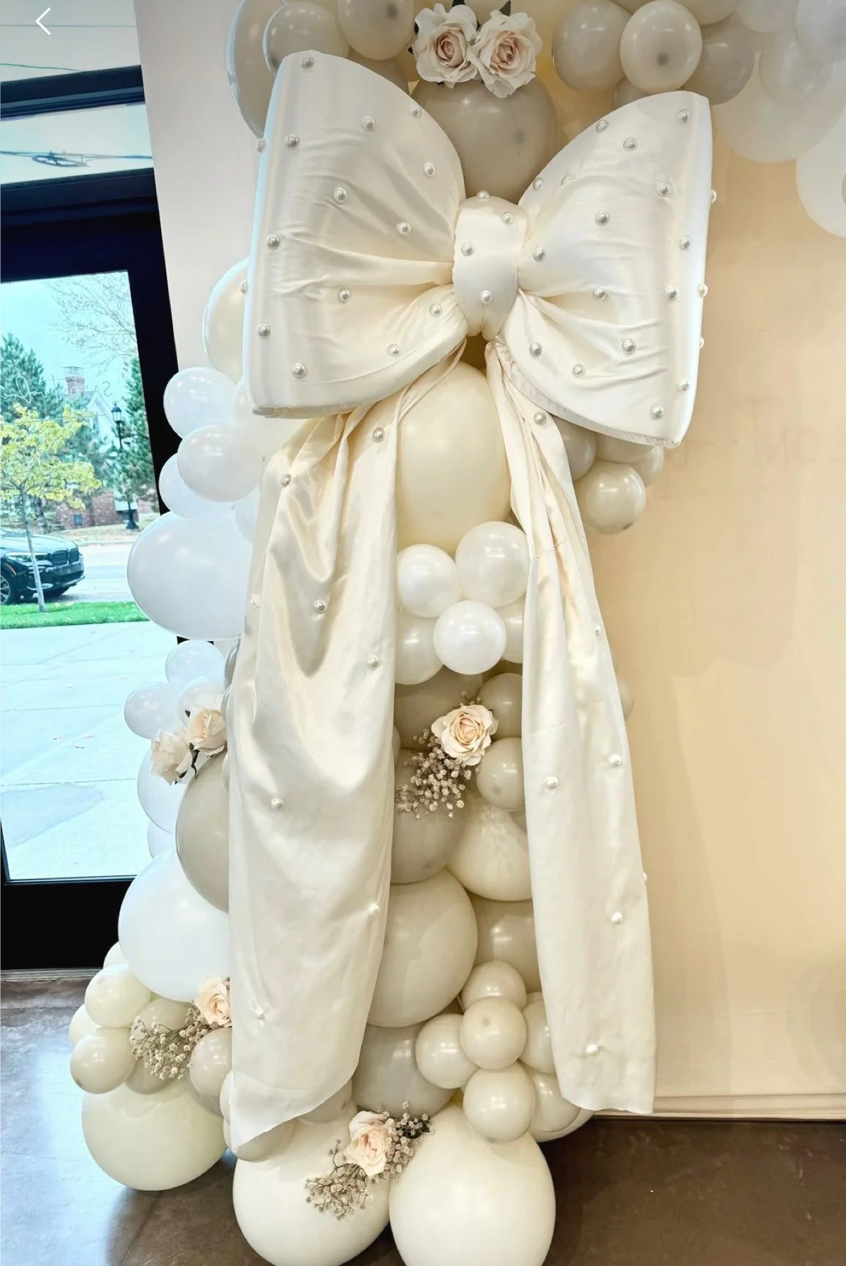 Decorative arrangement featuring a large white satin bow with pearl accents, surrounded by white balloons and roses, positioned near a window with a view of the street outside.