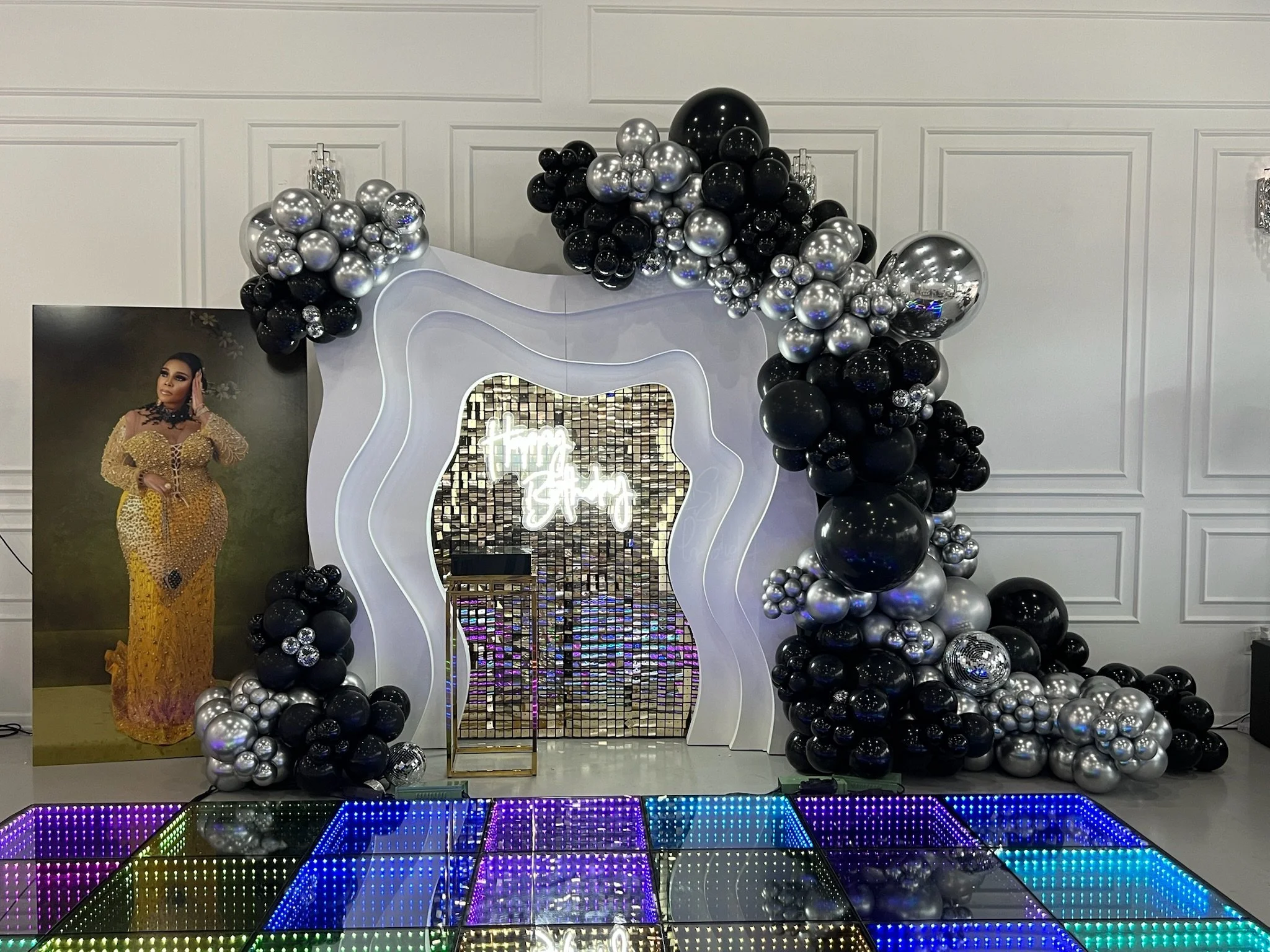 Decorative photo backdrop with black, silver, and mirrored balloons surrounding a neon sign, and a large poster of a woman in a yellow and gold gown on the left. Colorful lit floor tiles in front.