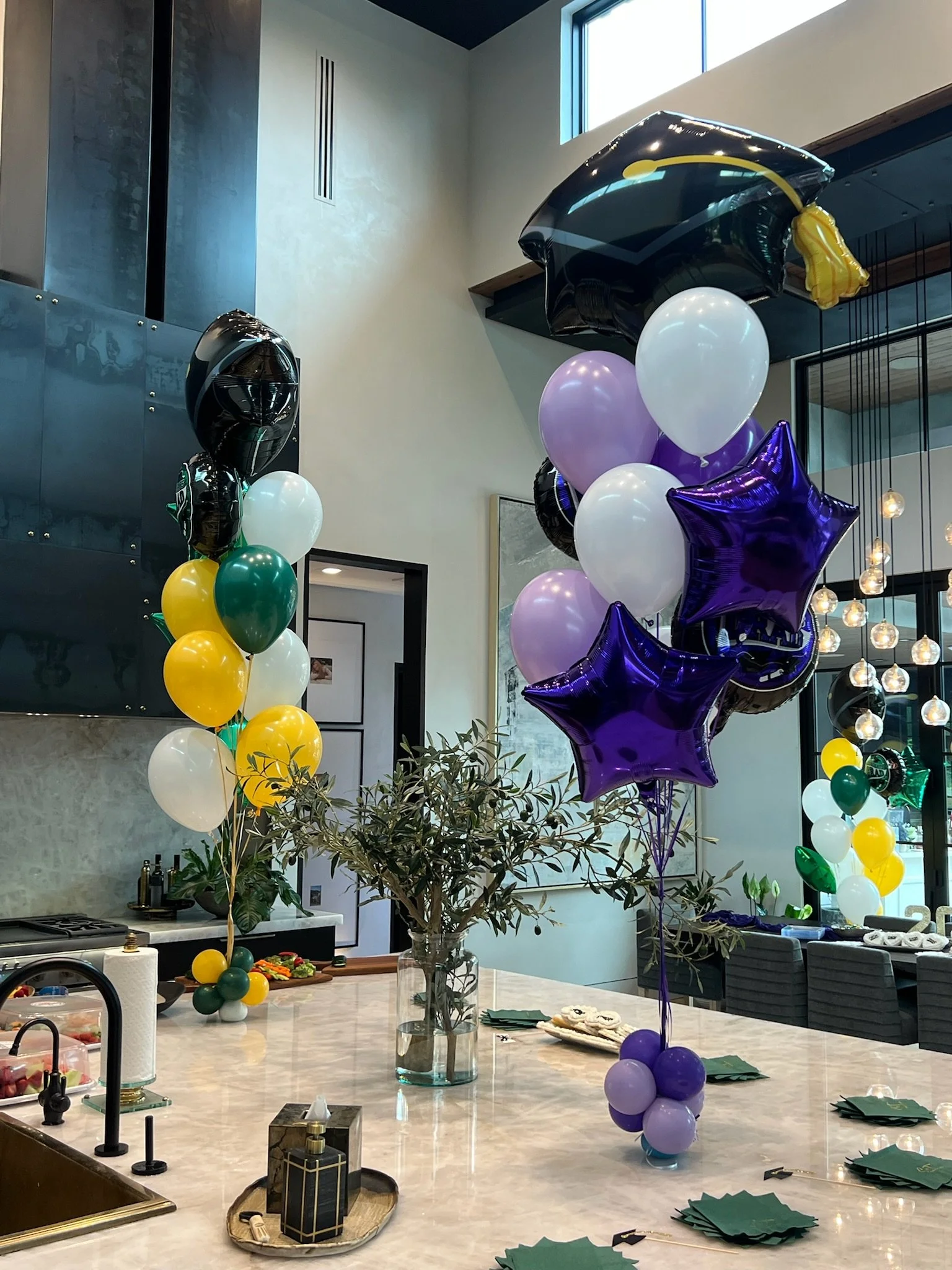 Decorative balloons on a kitchen island for a celebration, with a vase of greenery, napkins, and food in the background.