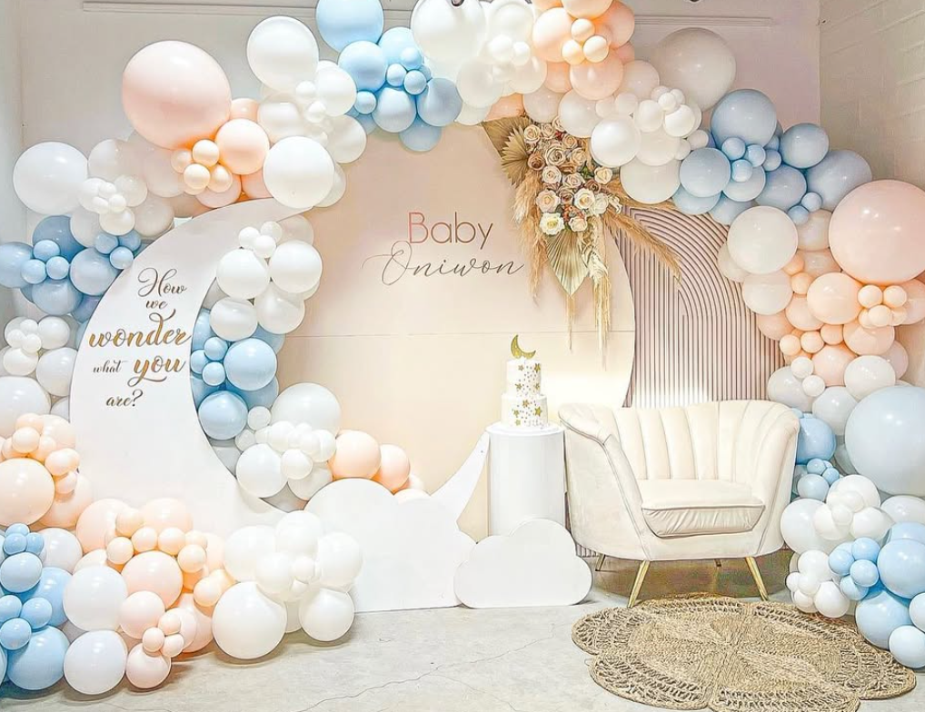 Decorative Baby Shower backdrop with pastel balloon arch in white, peach, and blue, featuring a crescent moon and clouds, a cake with a moon topper, and a plush cream armchair.