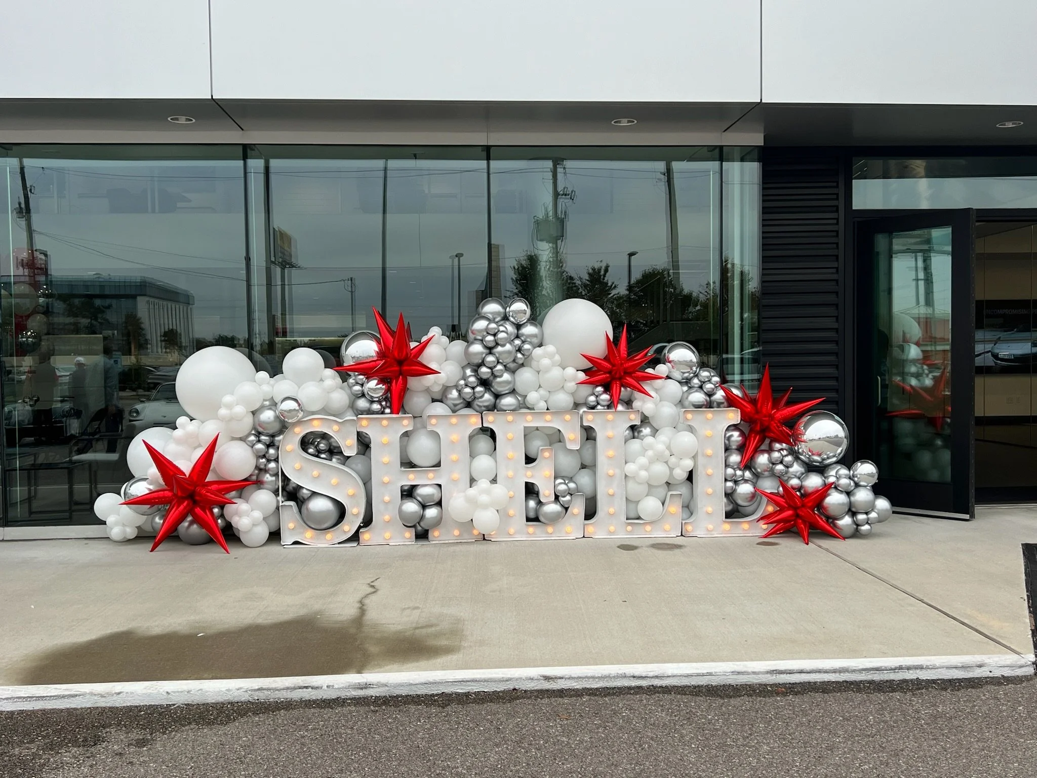 Holiday decoration with large illuminated letters spelling 'SALE' surrounded by red and silver balloons and stars outside a glass storefront.
