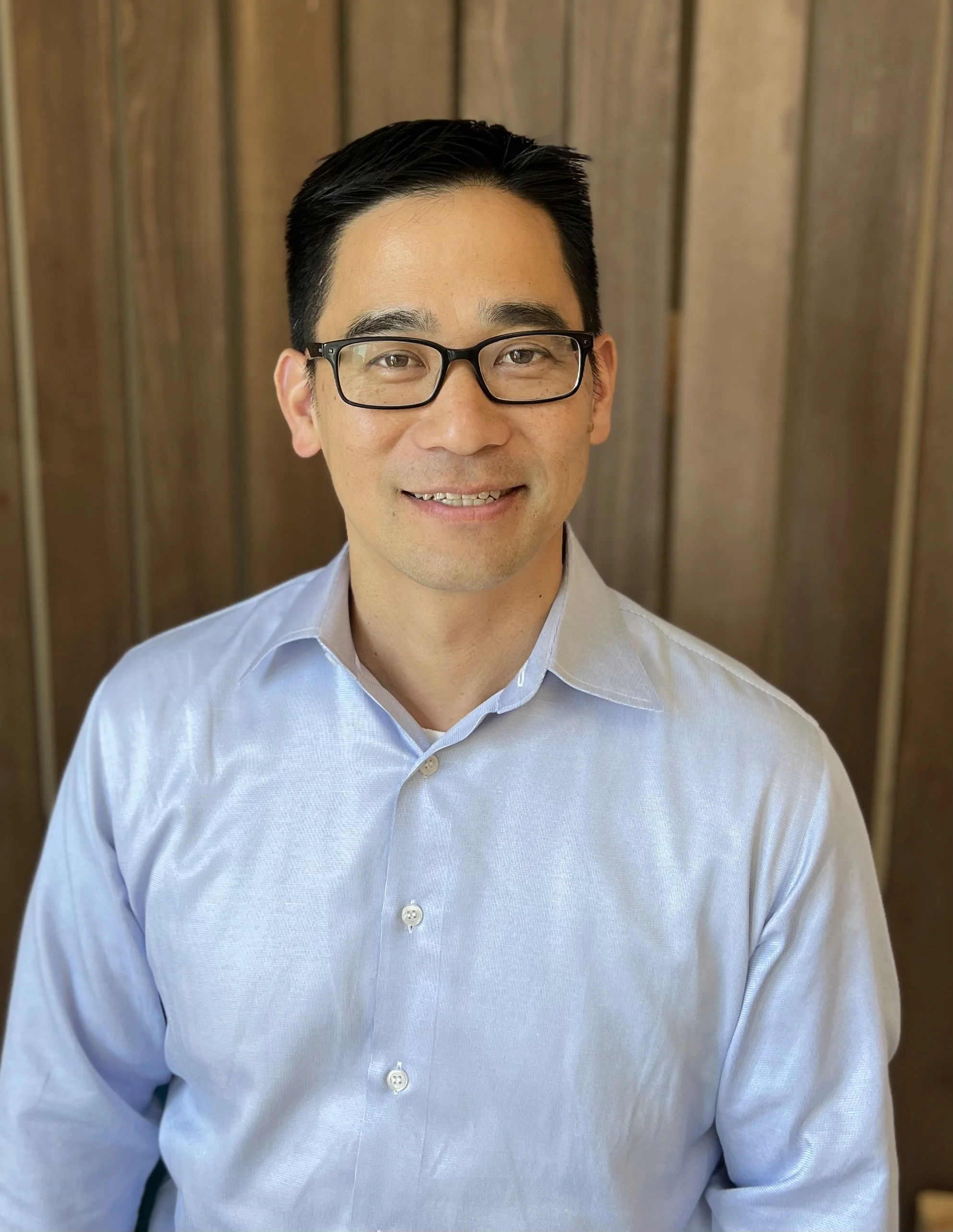 Board member with glasses wearing blue dress shirt smiling against wooden backdrop, Cocoon House Snohomish County