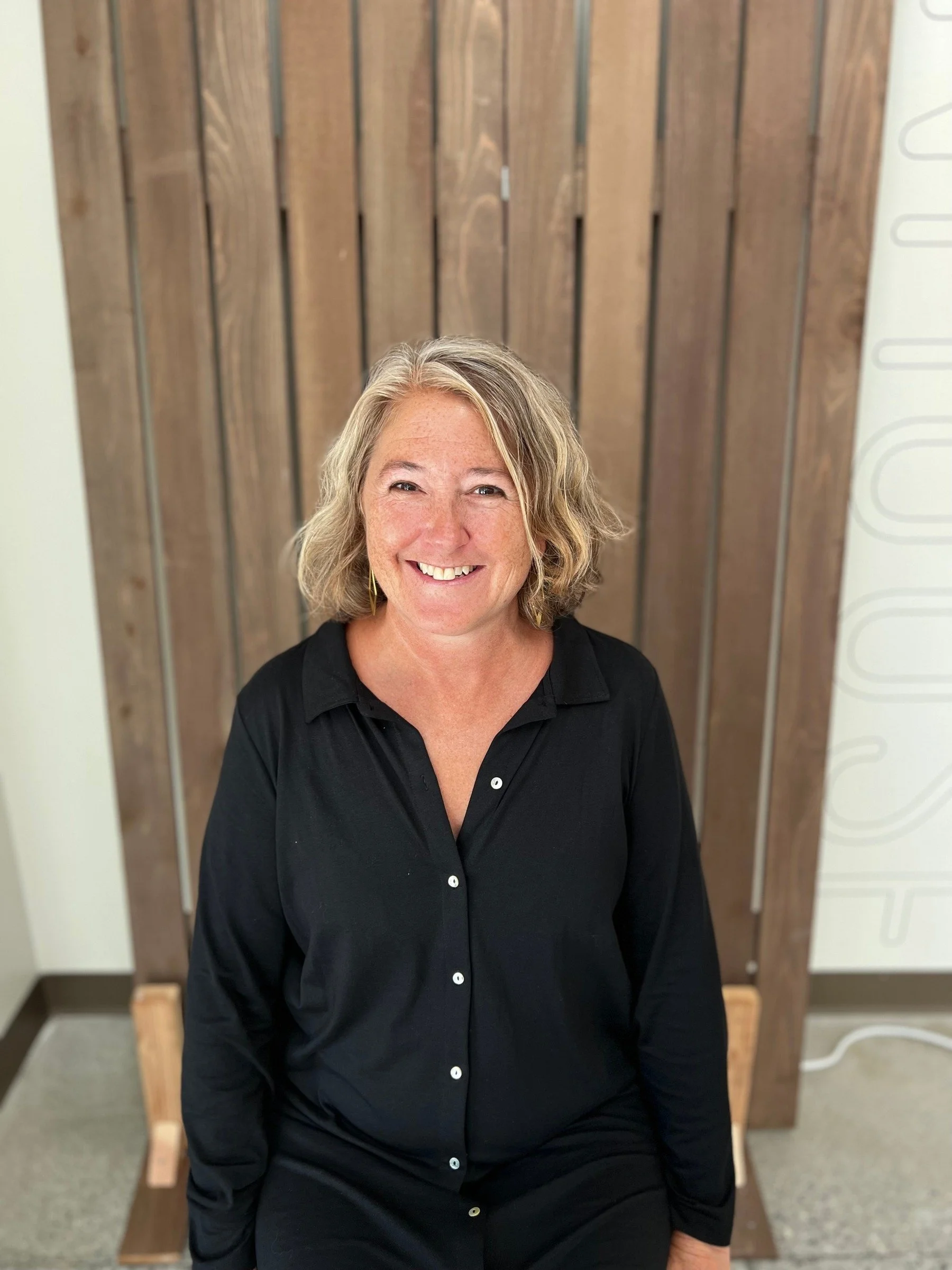 Board member with curly blonde hair smiling in seated portrait against wooden backdrop, Cocoon House Snohomish County