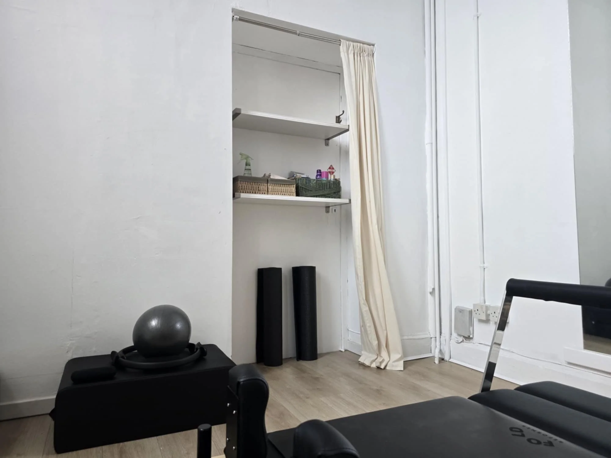 A fitness room with a black reformer machine, a medicine ball on a black pilates box, two black yoga mats, a white curtain, and shelves with gym accessories against a white wall.