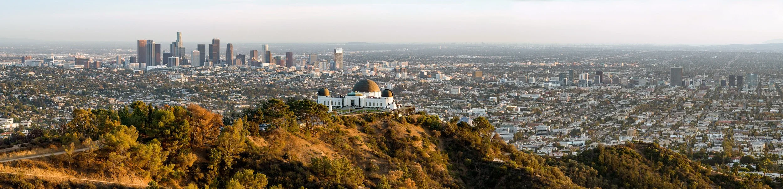 A panoramic cityscape view of Los Angeles at sunset, with the Griffith Observatory in the foreground on a hill, overlooking downtown LA and the surrounding city.