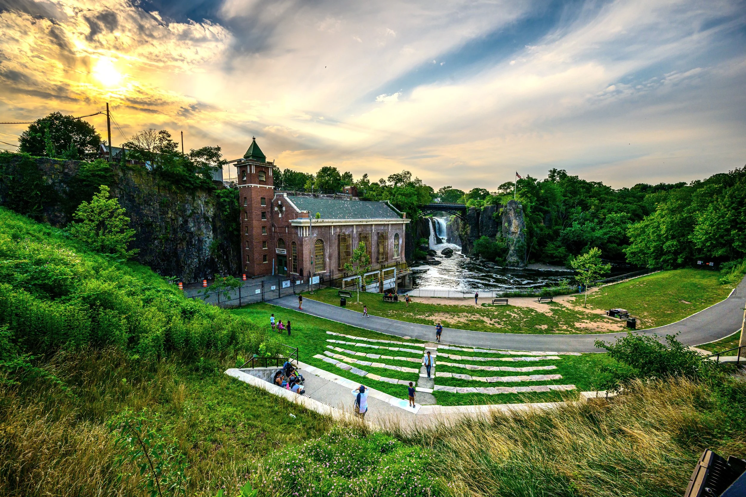 Scenic view of a park with a historic mill building near a waterfall, green trees, grassy slopes, and people walking and relaxing at sunset.