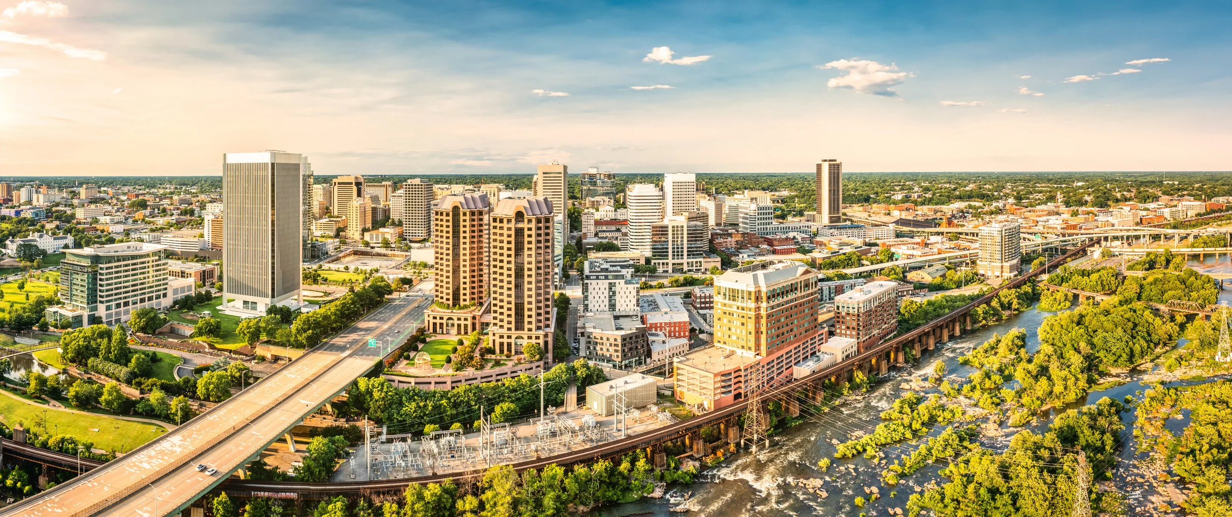 Aerial view of a city skyline with tall modern buildings, green parks, and a river with a bridge and railway tracks running through the city on a sunny day.