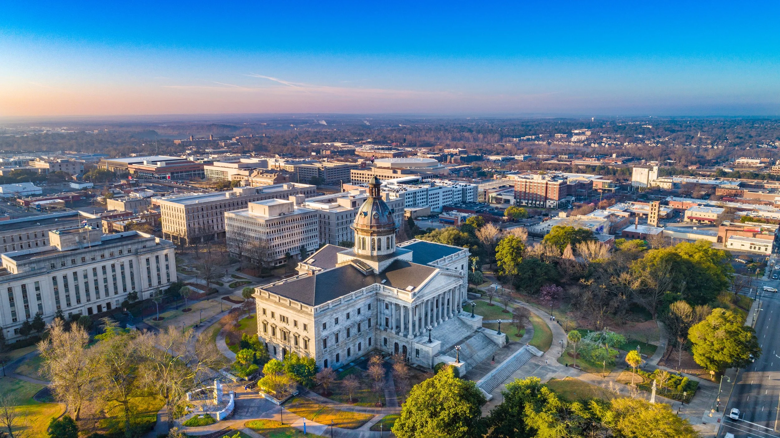 Aerial view of the capitol building in a city with surrounding trees and office buildings, under a clear blue sky.