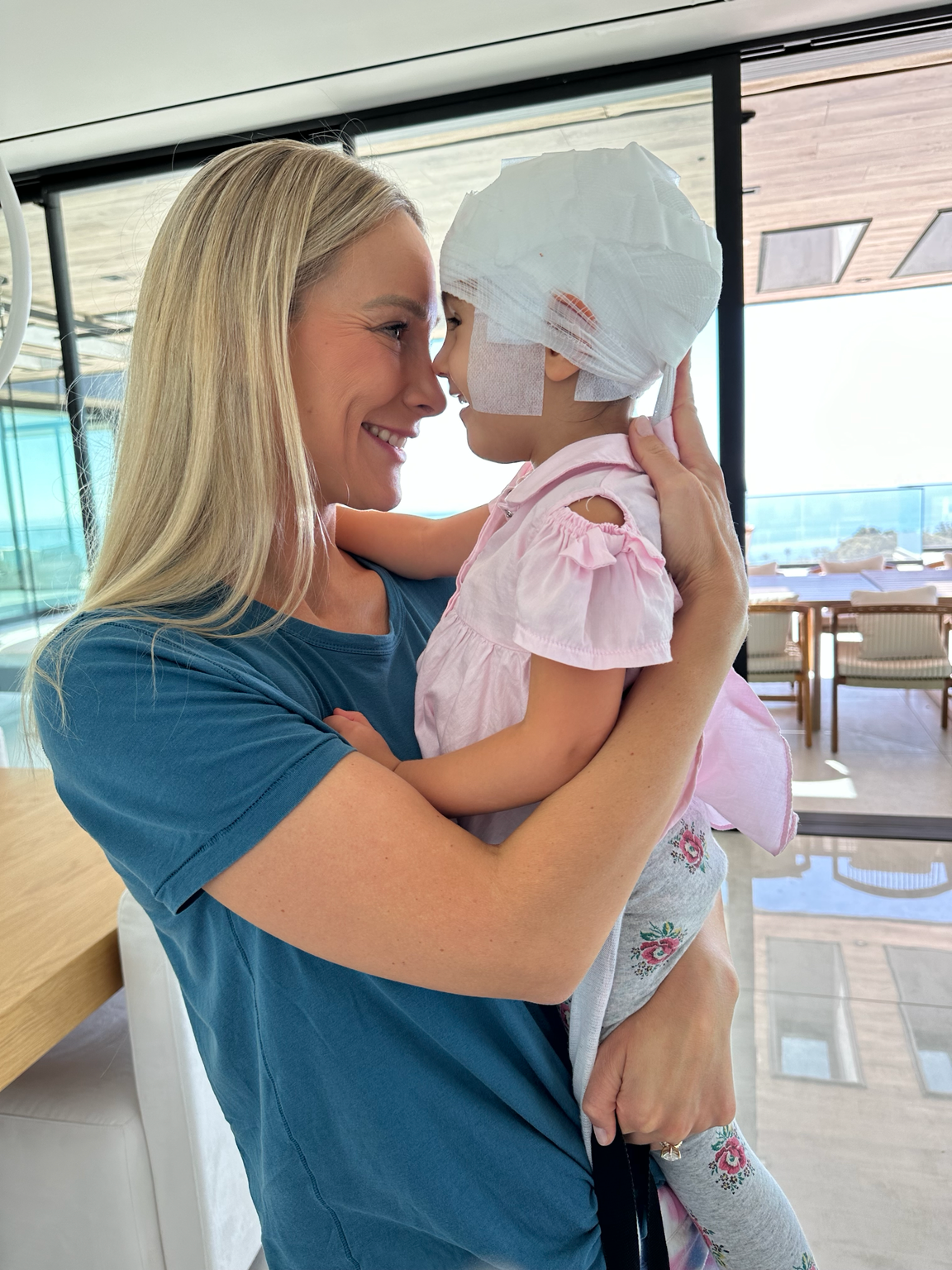 A woman holding a young girl with a bandaged head and face, both touching foreheads and smiling at each other in a bright indoor space.