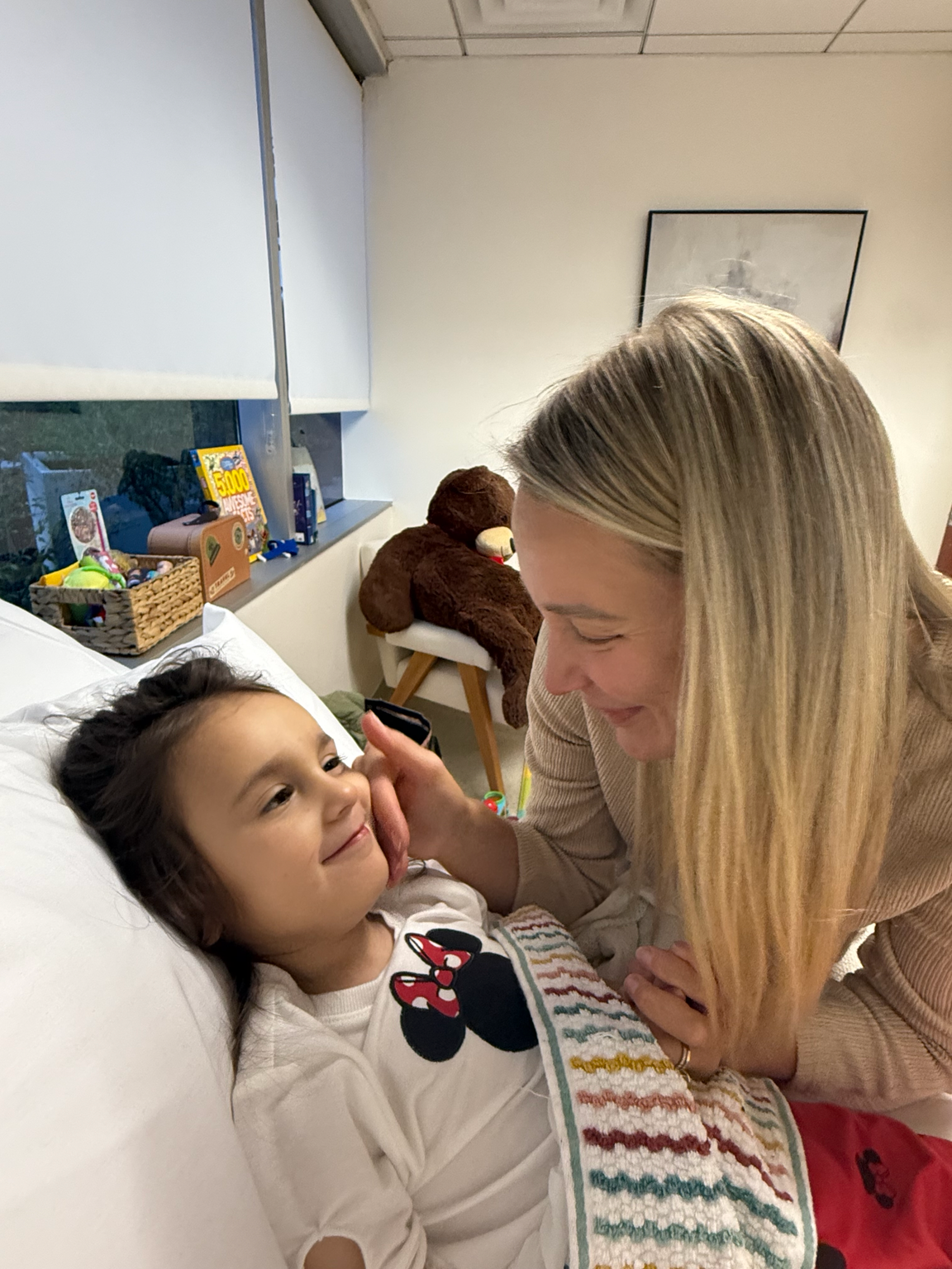 A young girl lying in a hospital bed, smiling, with a woman leaning over her and touching her face gently. The girl is wearing a white shirt with Minnie Mouse on it and a colorful blanket. In the background, there are stuffed animals, toys, and books on a shelf, with a large teddy bear sitting on a chair.