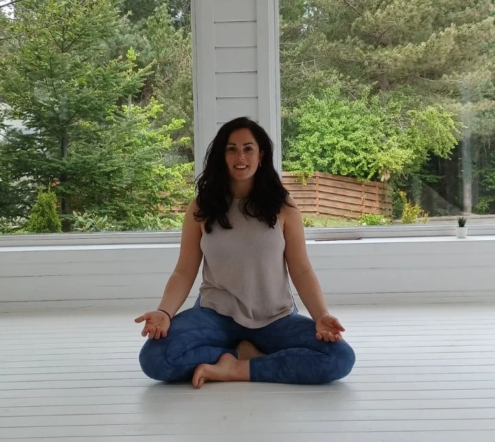 A woman practicing yoga indoors on a white floor with large windows behind her, showing green trees and a wooden fence outside.