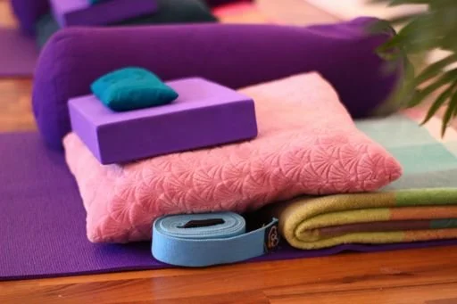 Yoga mat, folded towel, rolled yoga strap, and cushion on a wooden floor.