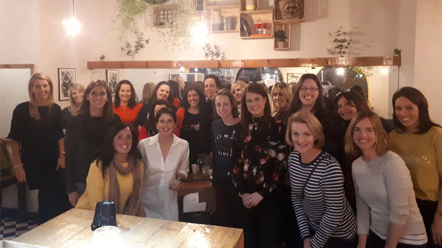 Group of women gathered together at a social event in a warmly lit room with wooden shelves and framed art on the wall.