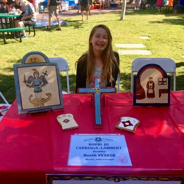 A young girl smiles behind a table displaying artwork.