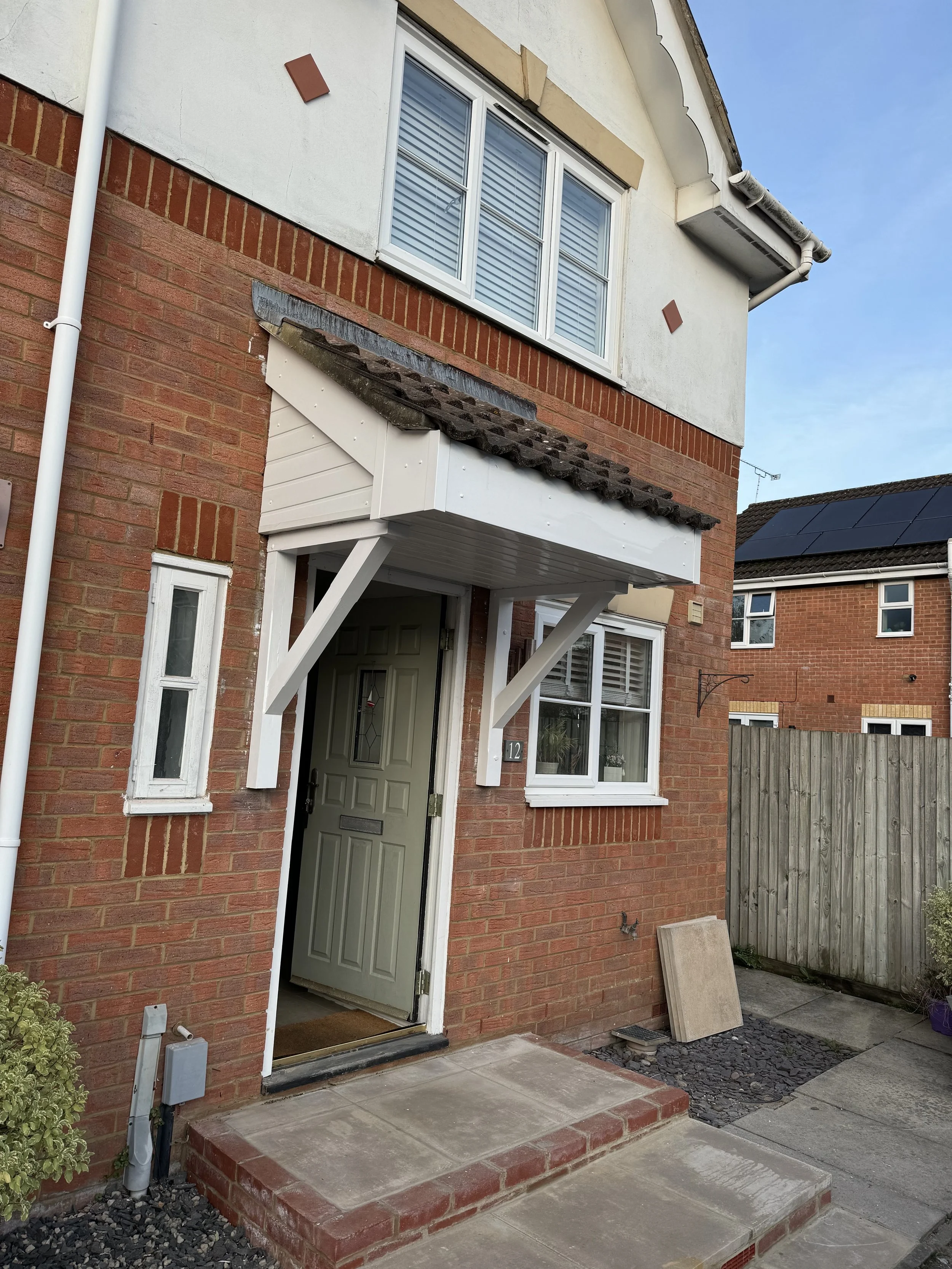 The front door of a two-story brick house with a small porch and white overhead cover. The house has white window frames and a small window next to the door. A wooden fence is visible to the right.