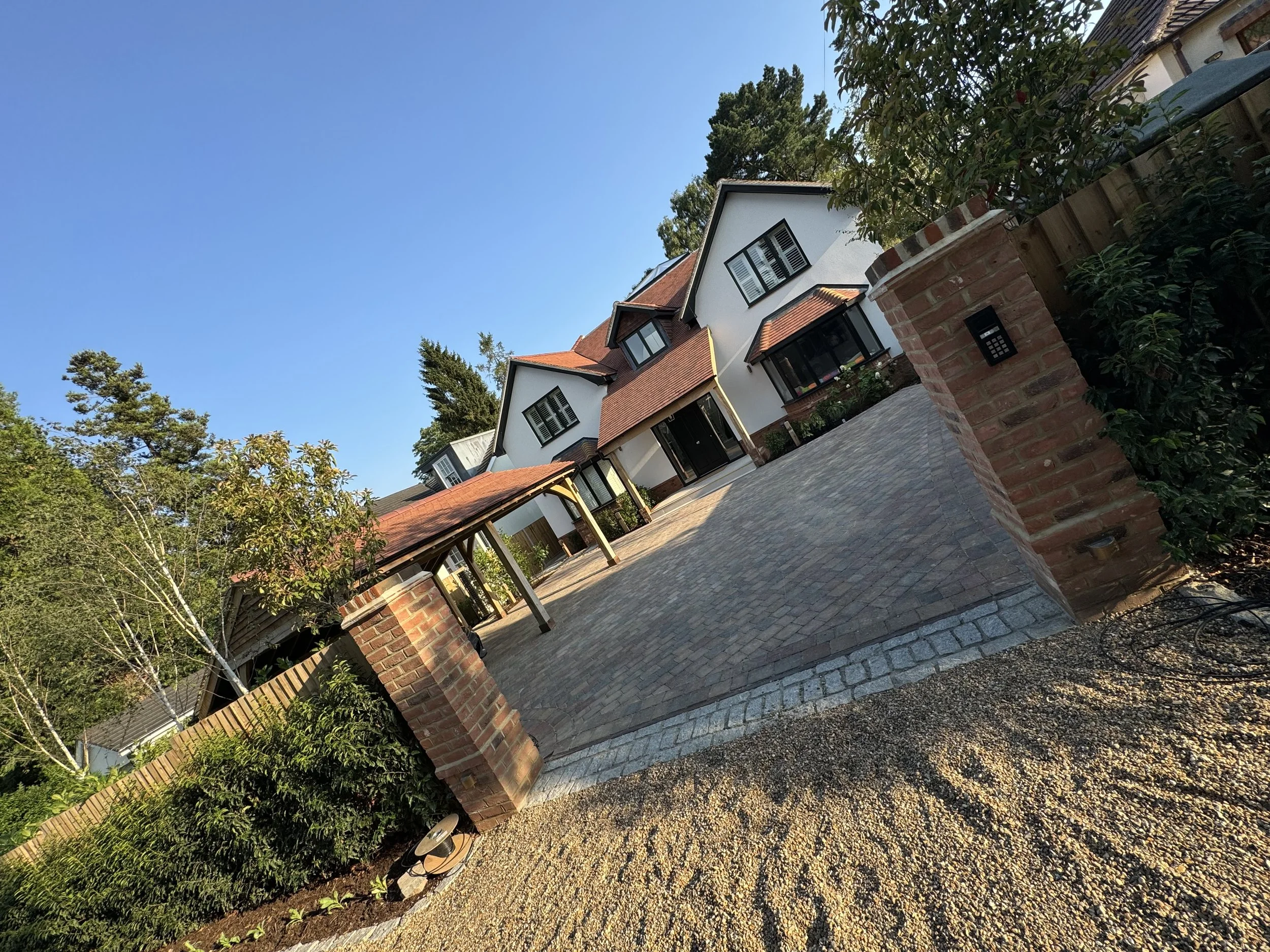 Modern multi-story house with white and brick exterior, surrounded by trees, under a clear blue sky, with a brick and pebble driveway, and a brick wall with a keypad entry system.