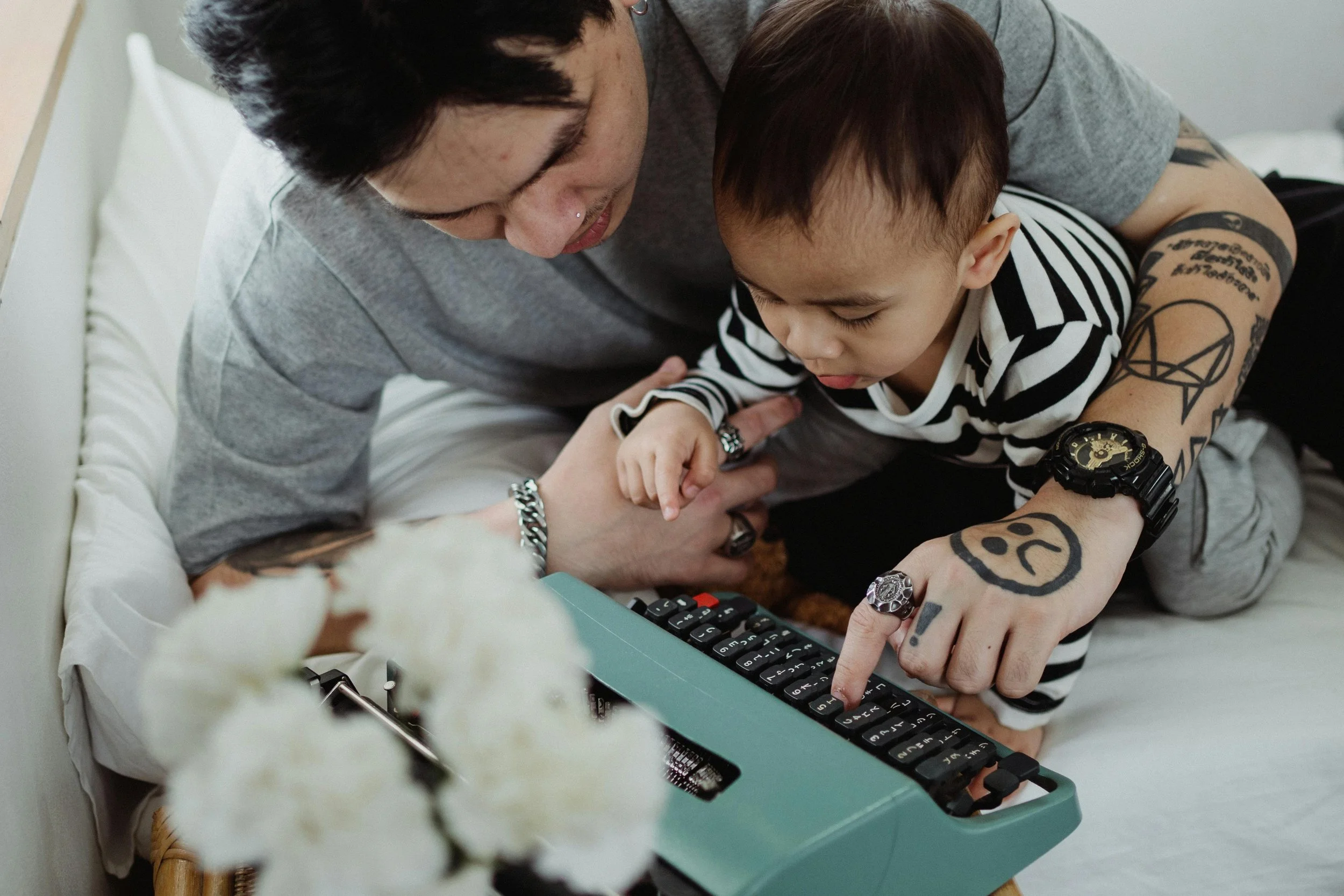 A man with tattoos and rings, wearing a gray shirt and a black watch, and a young boy with tattoos and rings, wearing a striped shirt, are on a bed looking at a vintage teal typewriter. The boy is pointing at the keys while the man holds his hand.
