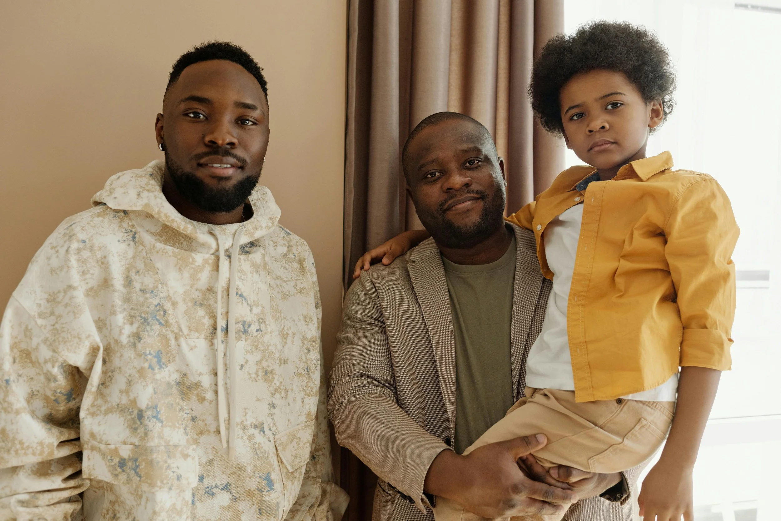 Three males, one child, standing indoors near beige curtains. The man on the left wears a light-colored, patterned hoodie. The man in the middle, dressed in a brown blazer and green shirt, holds a young boy with curly hair, wearing a yellow jacket, white shirt, and beige pants.