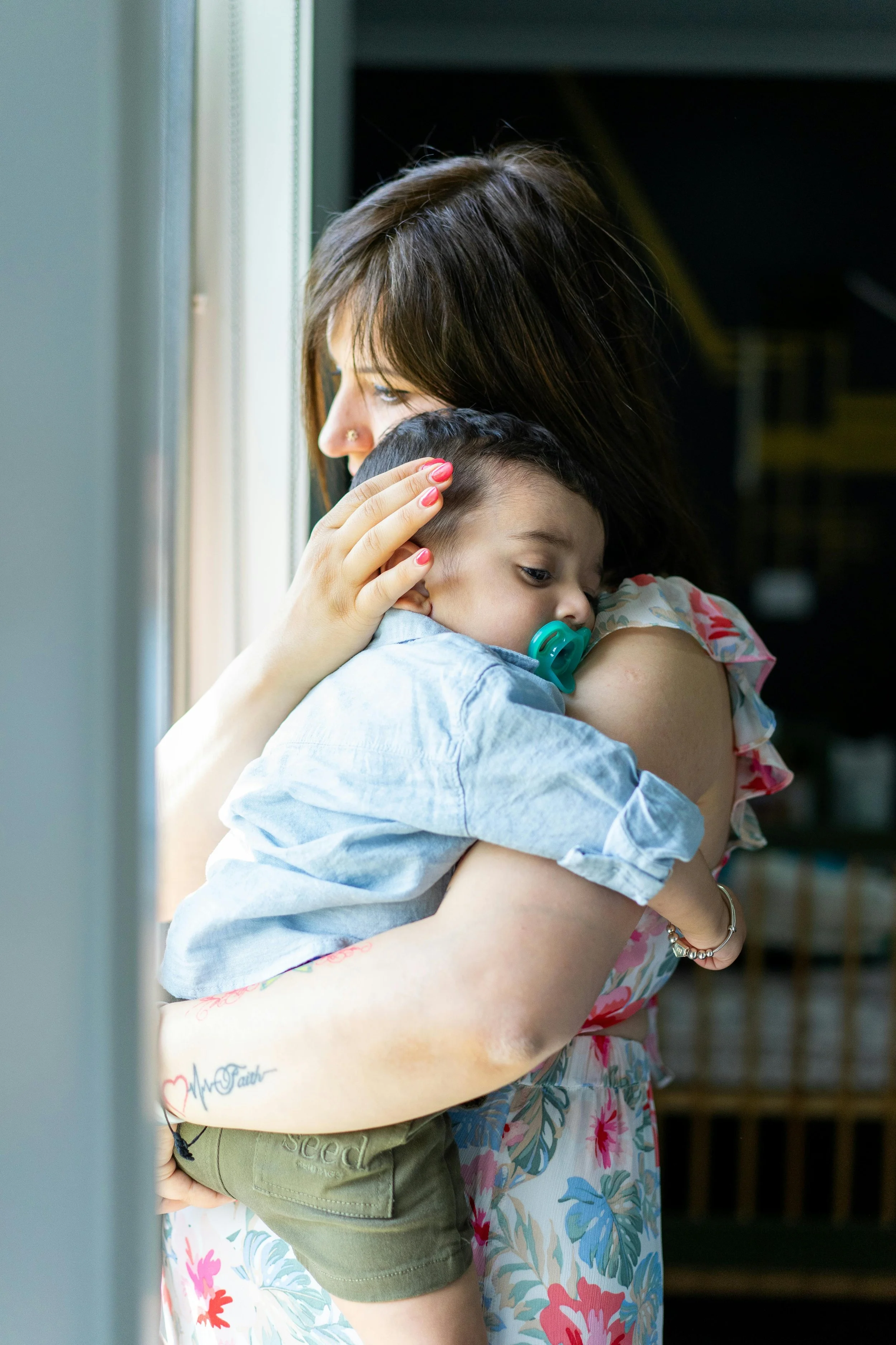 A woman with brown hair and a tattoo on her arm holding a young boy with a pacifier, looking out a window.