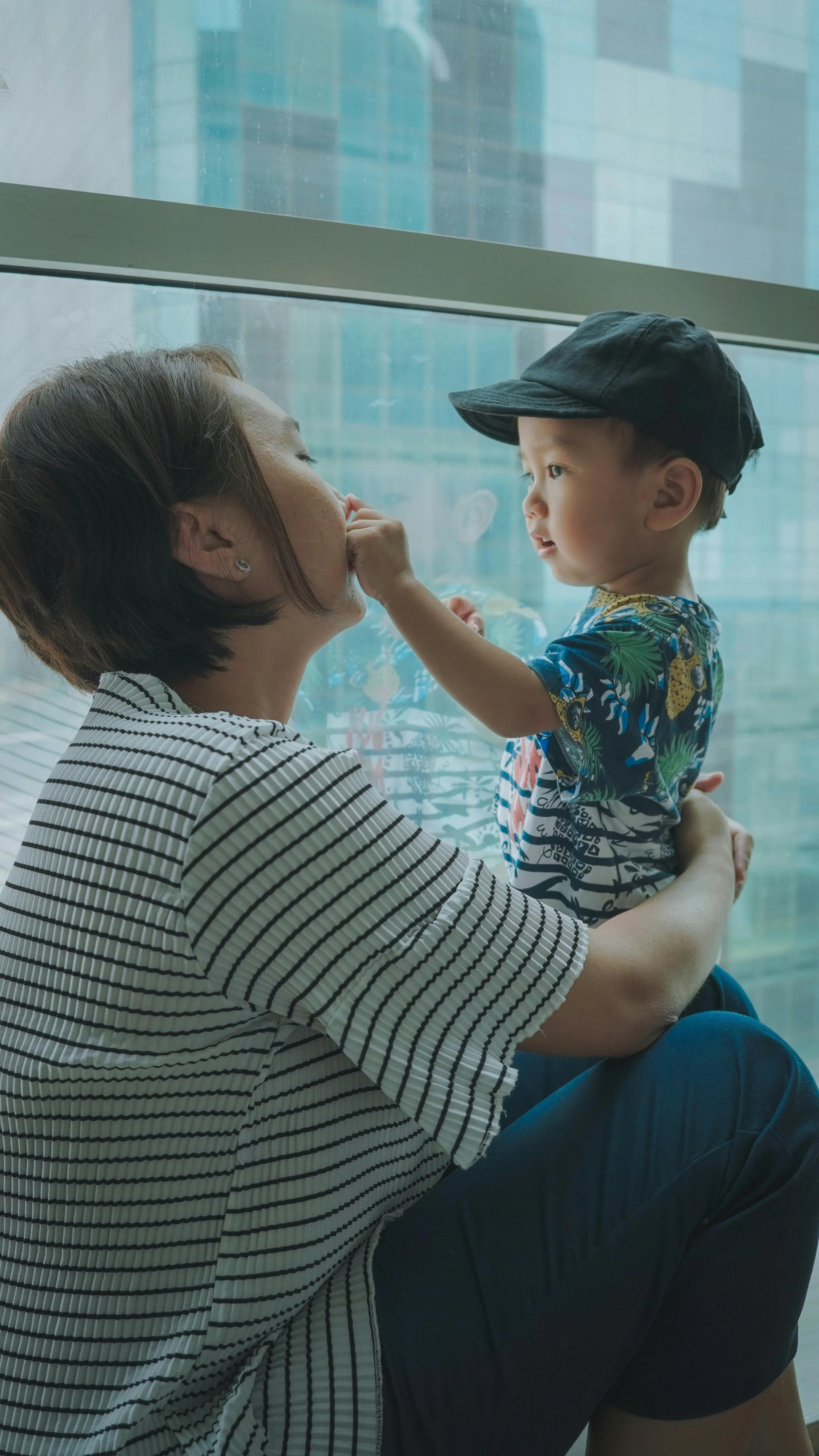 A woman and a young boy sitting by a large window. The woman is leaning in with her eyes closed, while the boy gently touches her nose. The boy wears a black cap and a colorful shirt with a tropical pattern.
