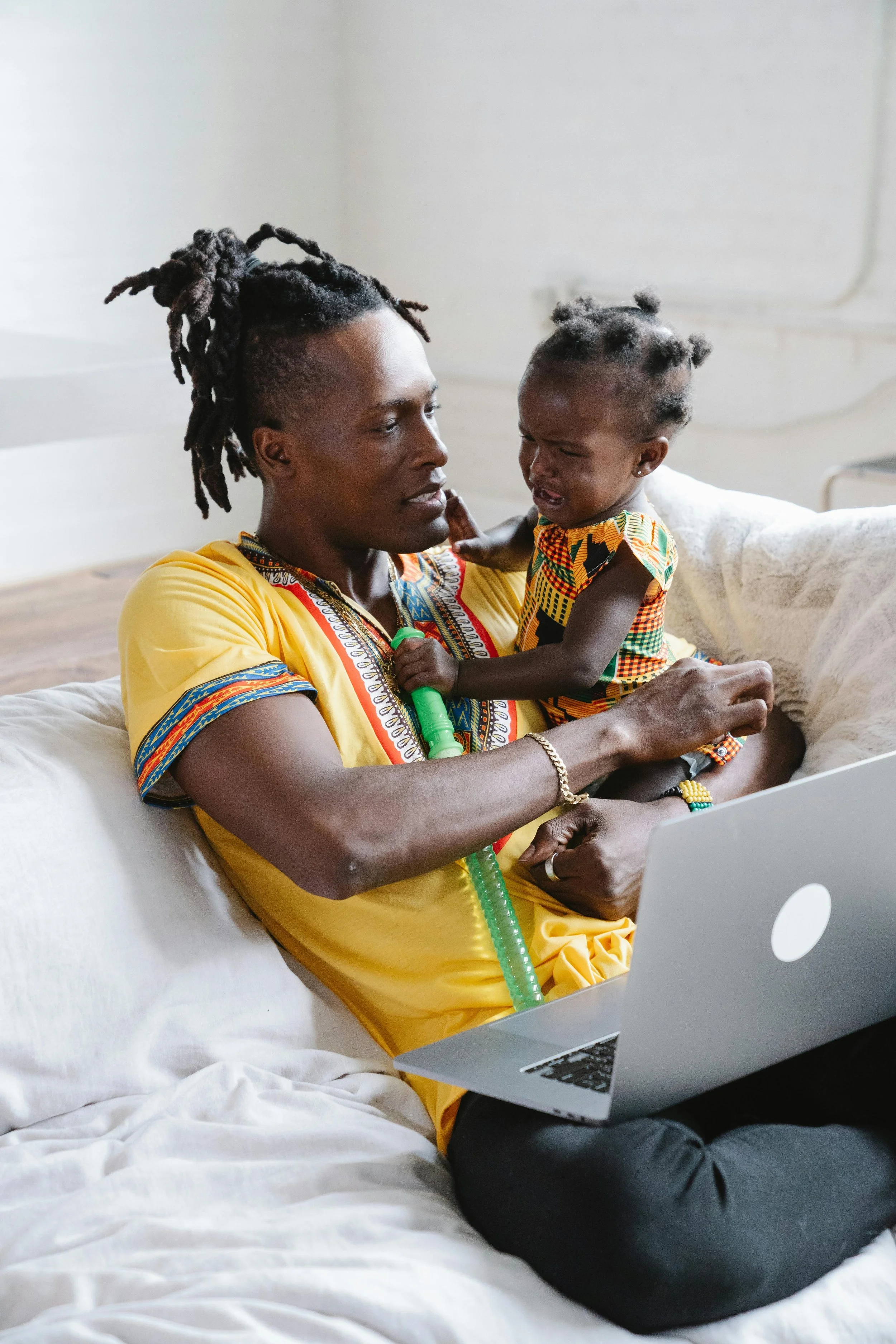 A woman wearing traditional African clothing holding a crying young child while sitting on a bed, using a laptop.