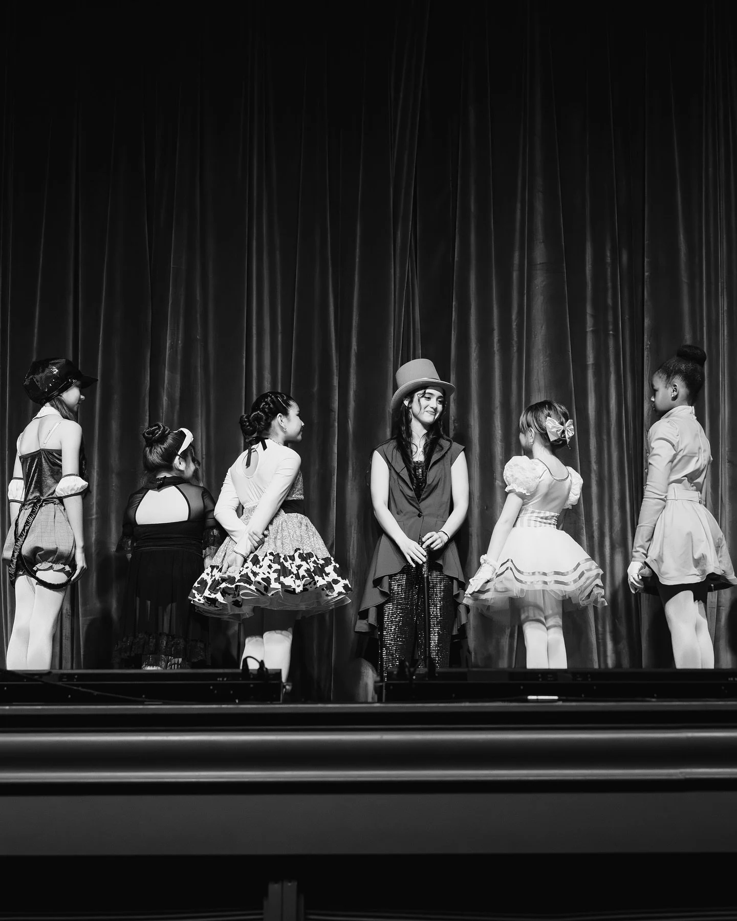 Black and white photo of a group of young girls on stage, dressed in various costumes, with one girl in the center wearing a top hat and a dark vest, smiling, against a backdrop of curtains.