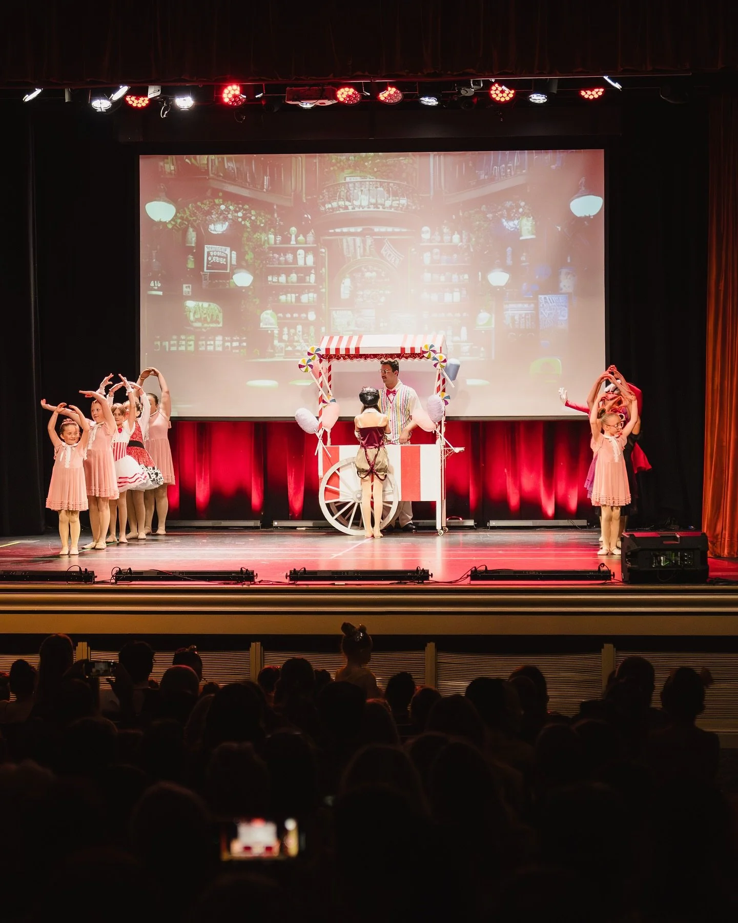 Children's theatrical performance on stage with a candy-themed cart and backdrop of a cozy cafe scene.