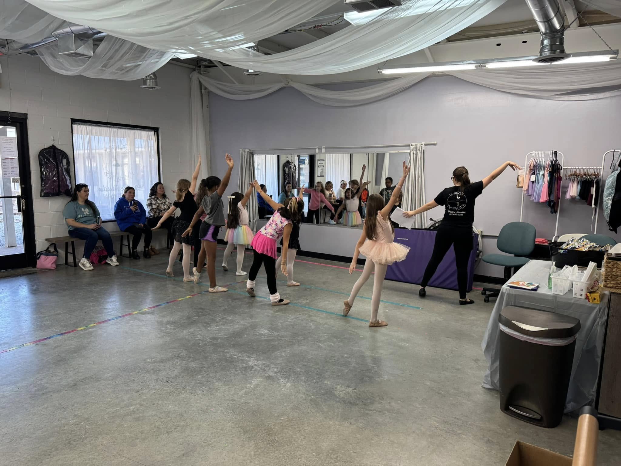 Ballet class with young girls stretching and practicing dance moves in studio, some sitting on bench and others watching in mirrors, instructor guiding students, dancewear hanging on racks.