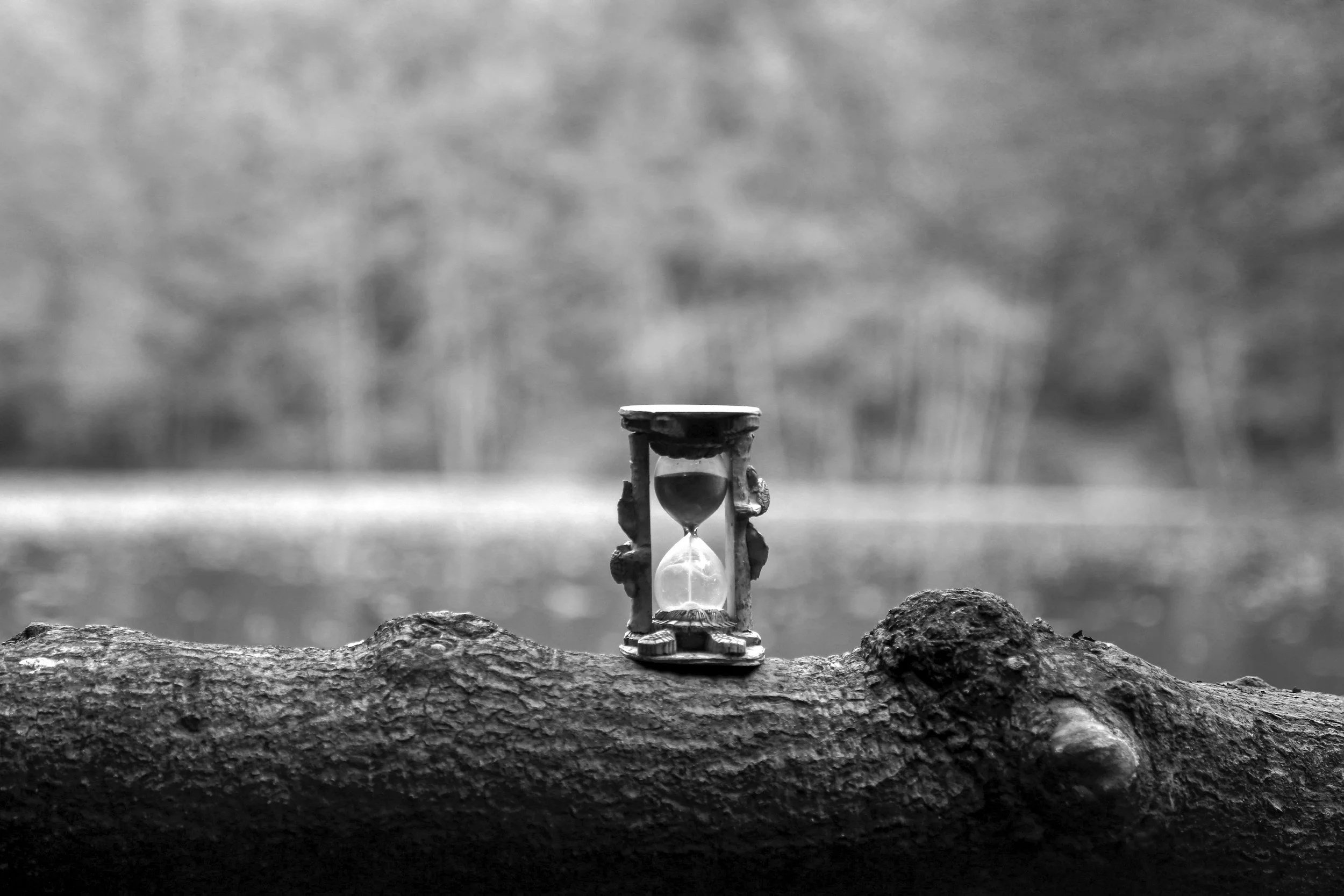 A small decorative hourglass with ornate legs and a detailed top sits on a rough surface, with a blurred background in grayscale.