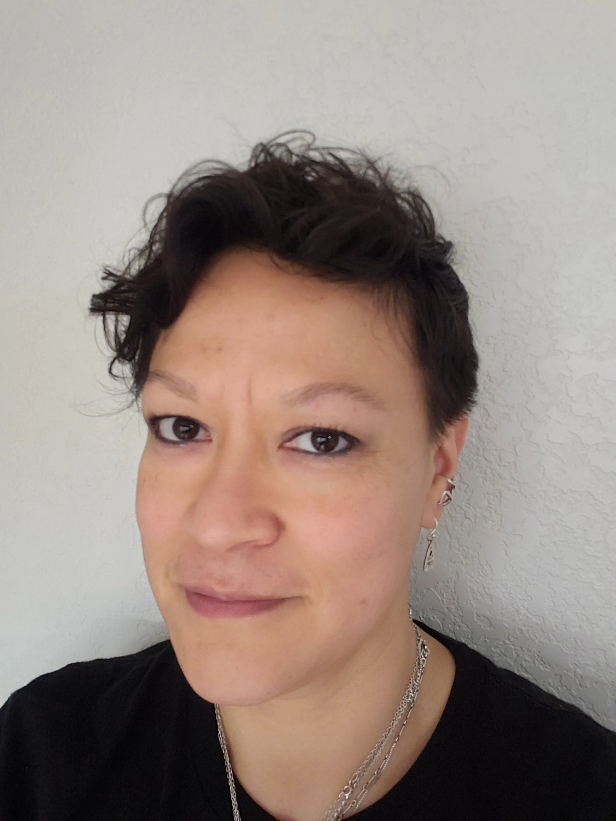 A person with short, dark hair, brown eyes, wearing a black shirt, silver jewelry, and earrings, standing against a plain light-colored wall.