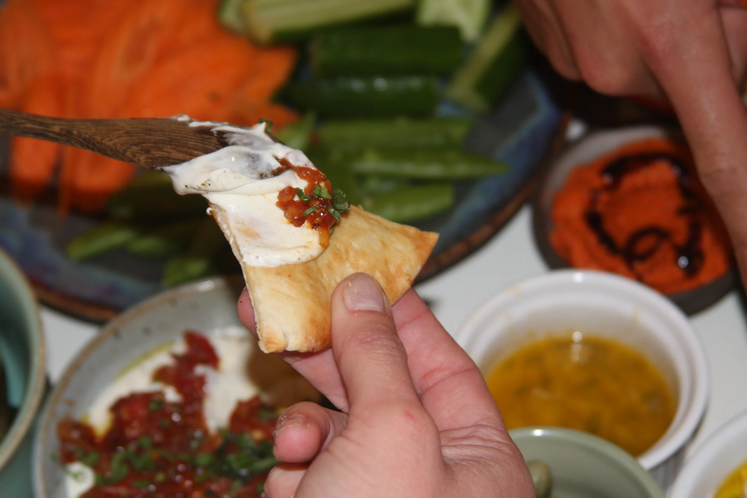 Hand holding a small piece of bread with sour cream, salsa, and chives, with a background of various dishes including fresh vegetables, salsa, and dips.