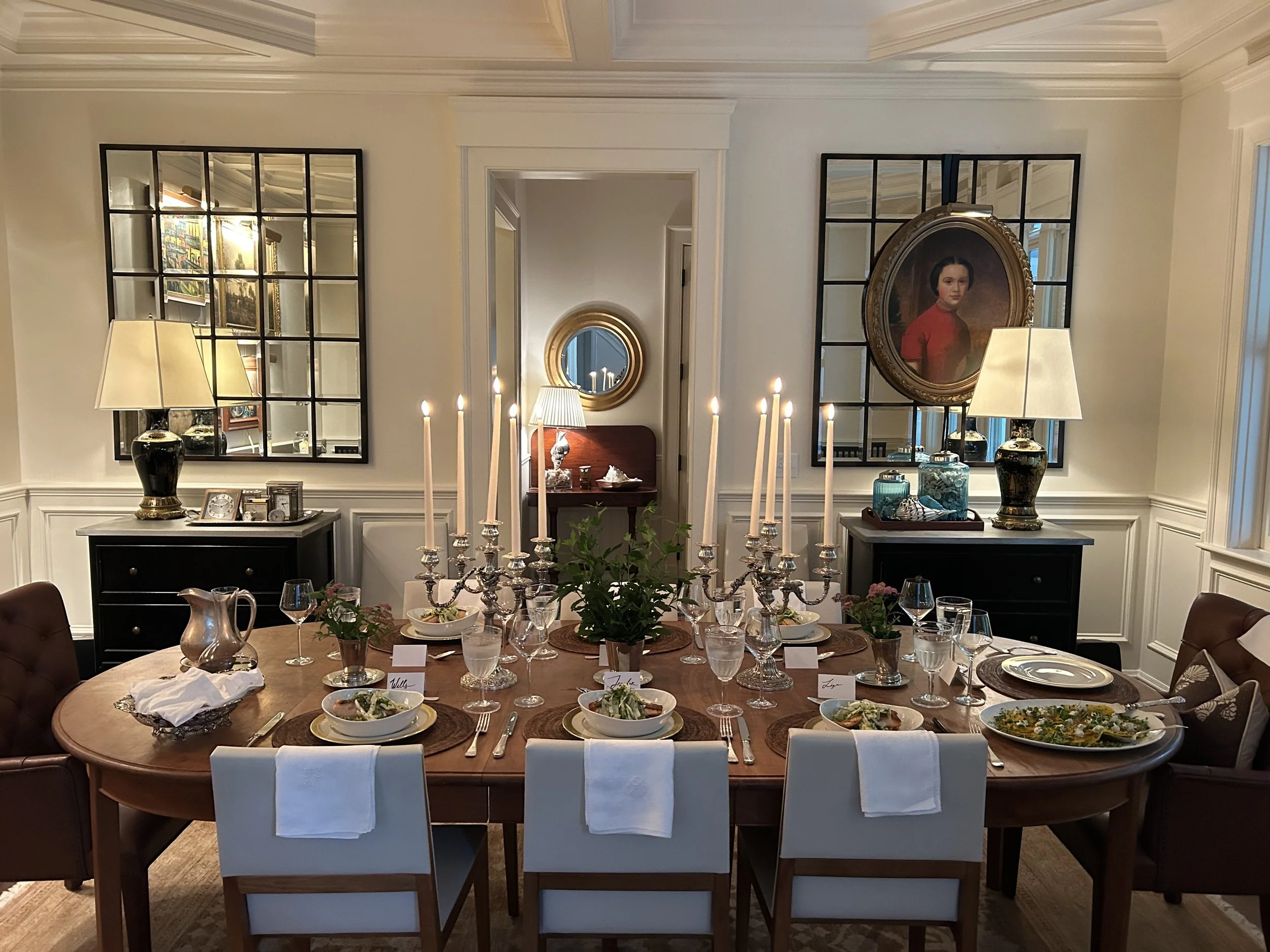 A dining room with a large oval table set for a meal, featuring candles, dishes, and glasses. There are two black sideboards with lamps and decorative items, and framed mirrors and a portrait on the walls.