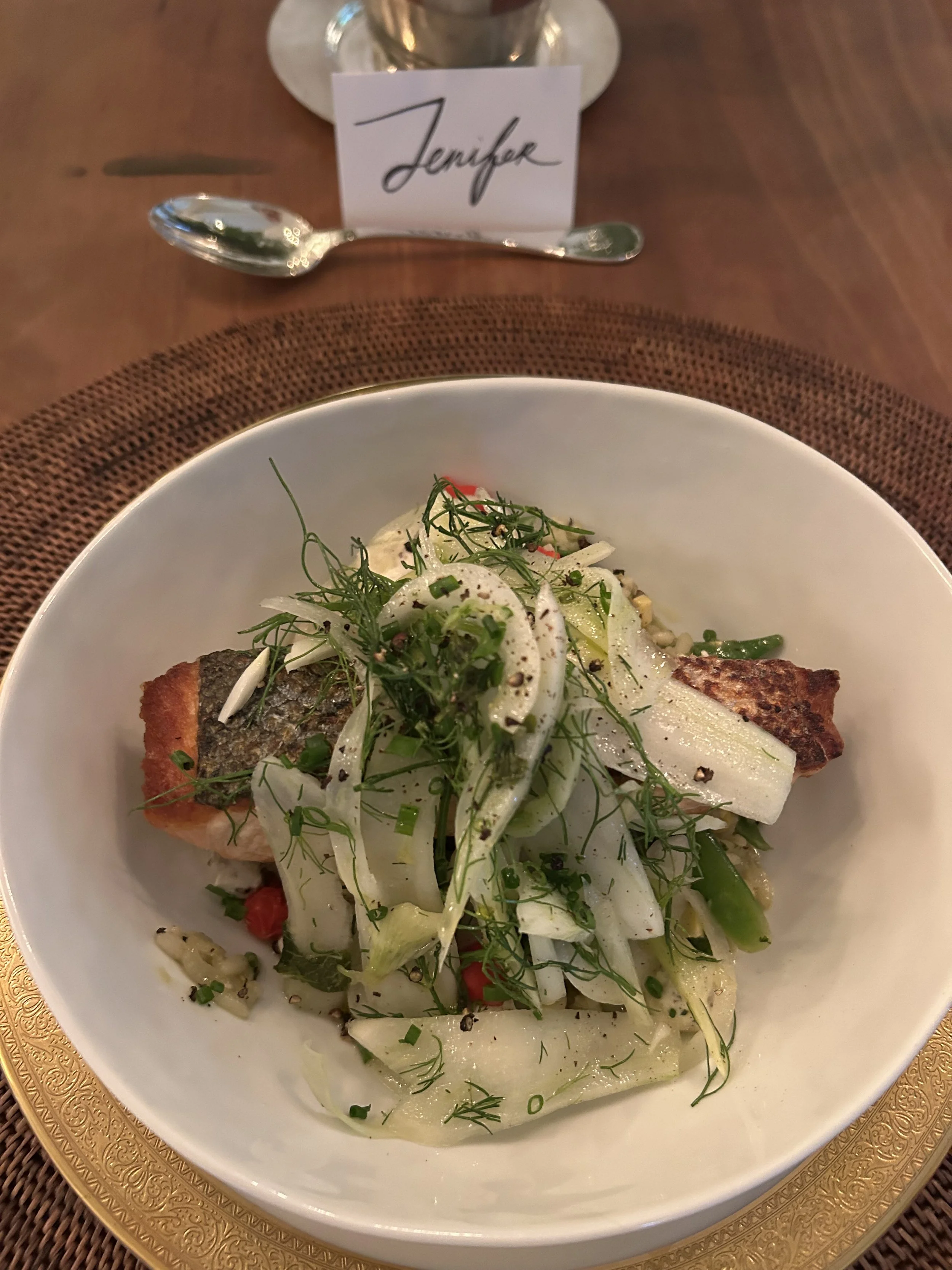 A white bowl filled with grilled fish and a fennel salad garnished with herbs and black pepper, placed on a gold-rimmed charger plate on a wooden table.