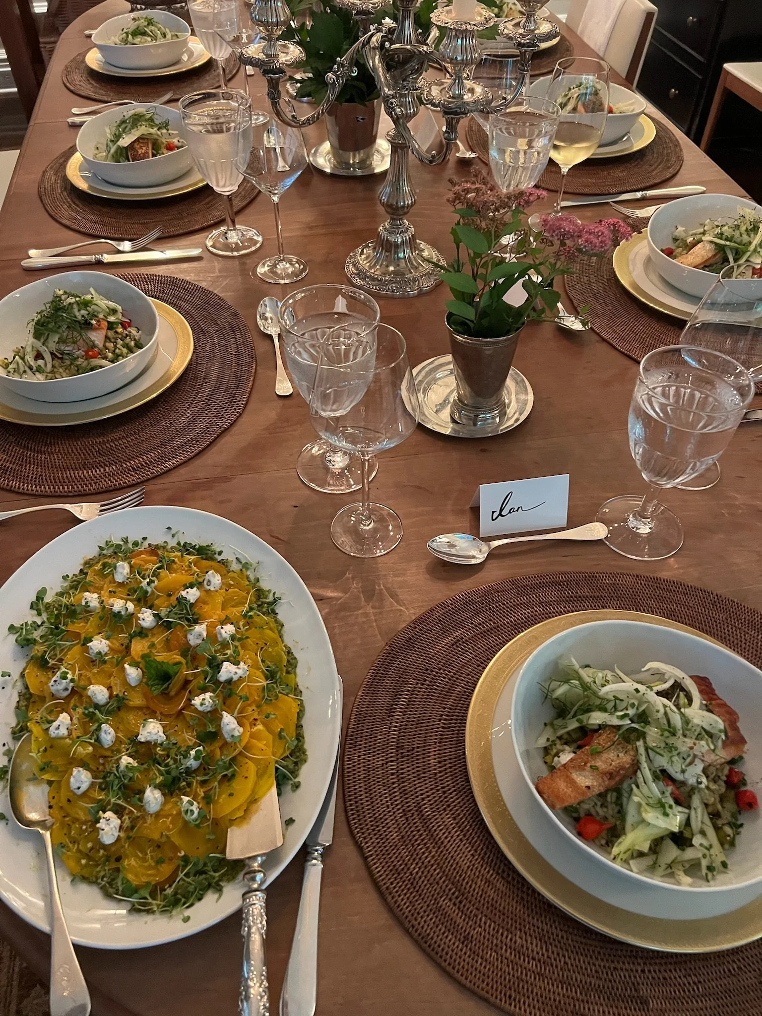 A formal dinner table set with plates of salad and pasta, wine glasses, water glasses, silverware, a silver candelabra, and a potted flower centerpiece.