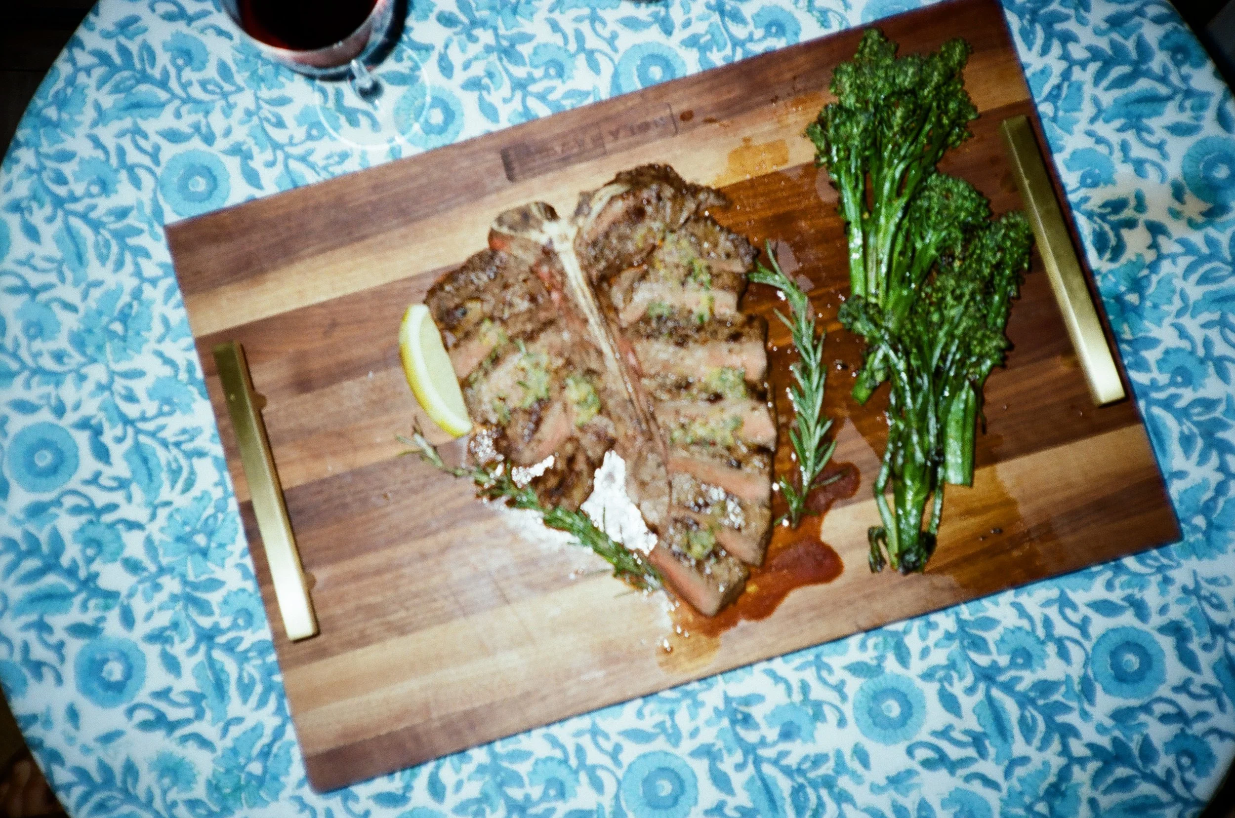 A cooked steak served on a wooden platter with lemon wedges, fresh herbs, and green broccolini on a patterned blue and white tablecloth.