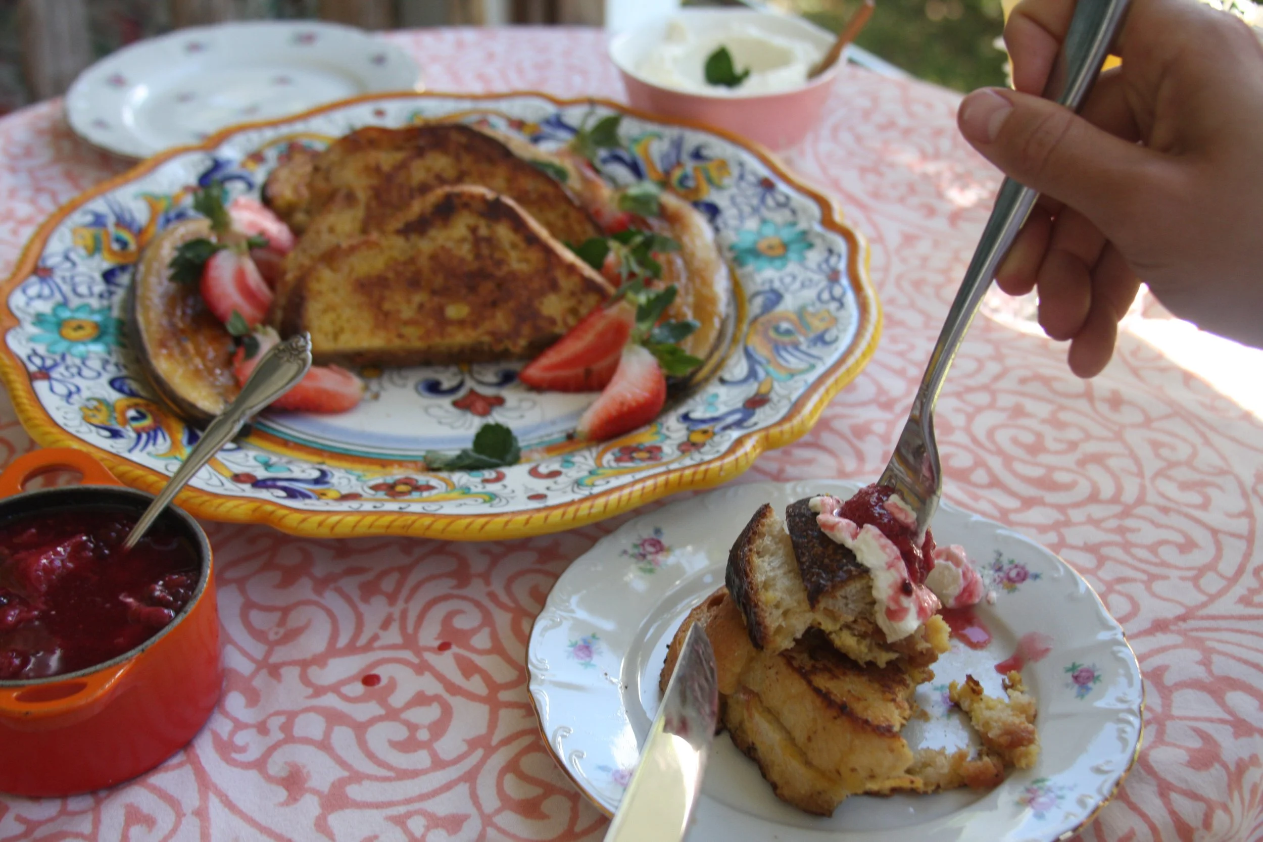 A table set with a large decorative plate of French toast with strawberries and mint garnishes, a small bowl of whipped cream, a small orange dish of berry compote, and a plate with a slice of French toast being cut with a fork, all on a pink pattern
