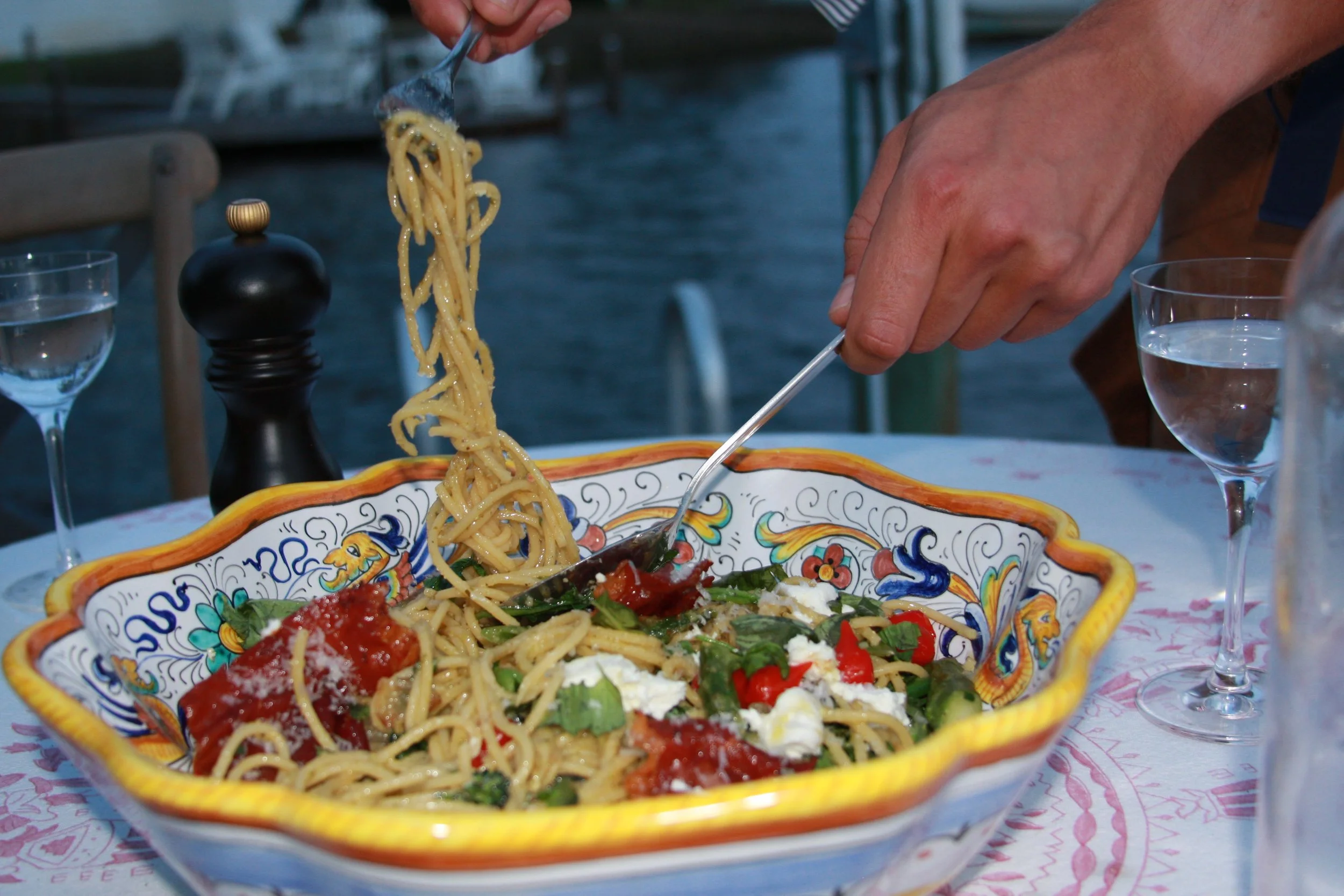 Person serving spaghetti with tomato sauce and vegetables from a decorative ceramic bowl at an outdoor restaurant table.