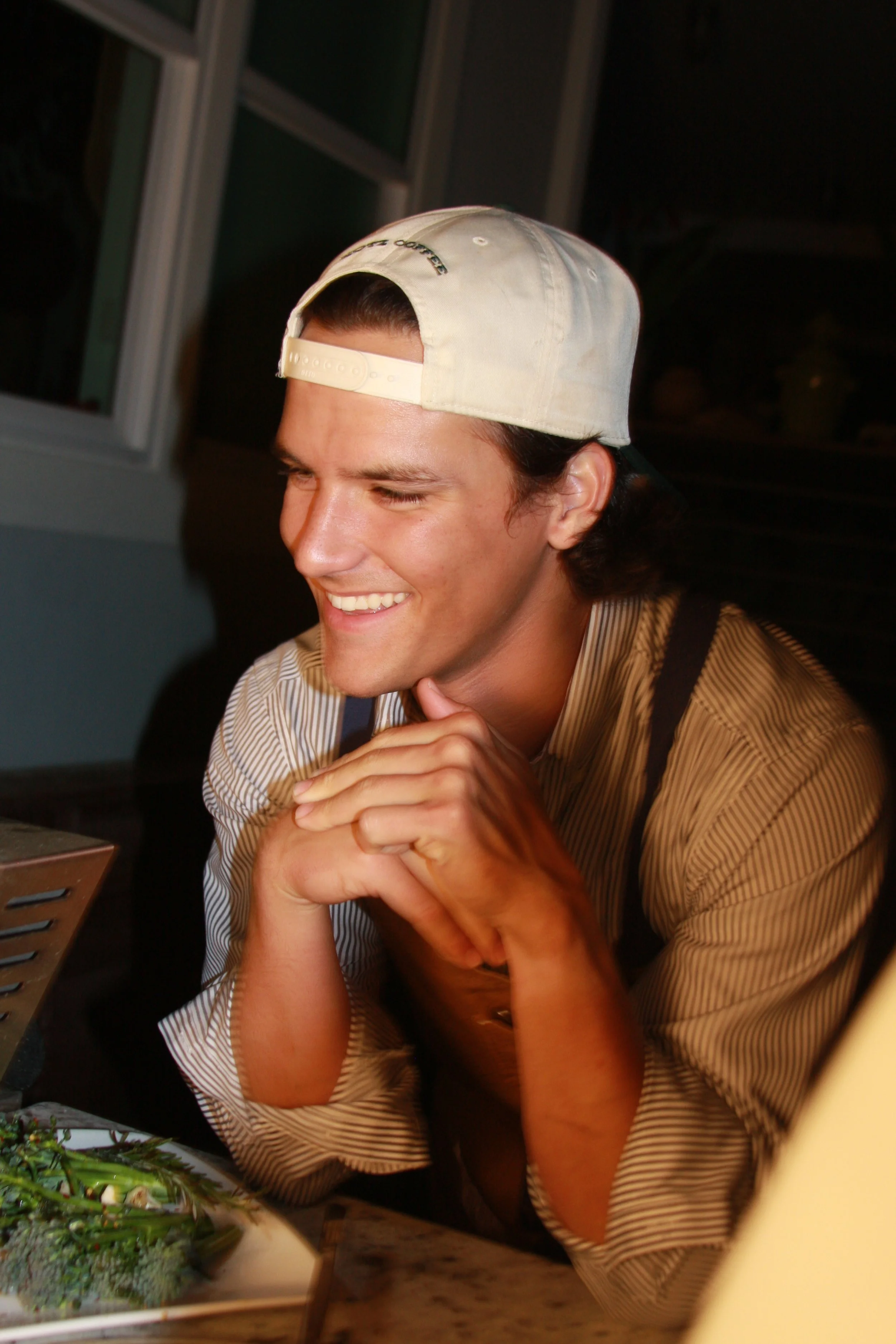 A young man smiling and sitting at a table with a plate of food, wearing a backward baseball cap and a striped shirt.