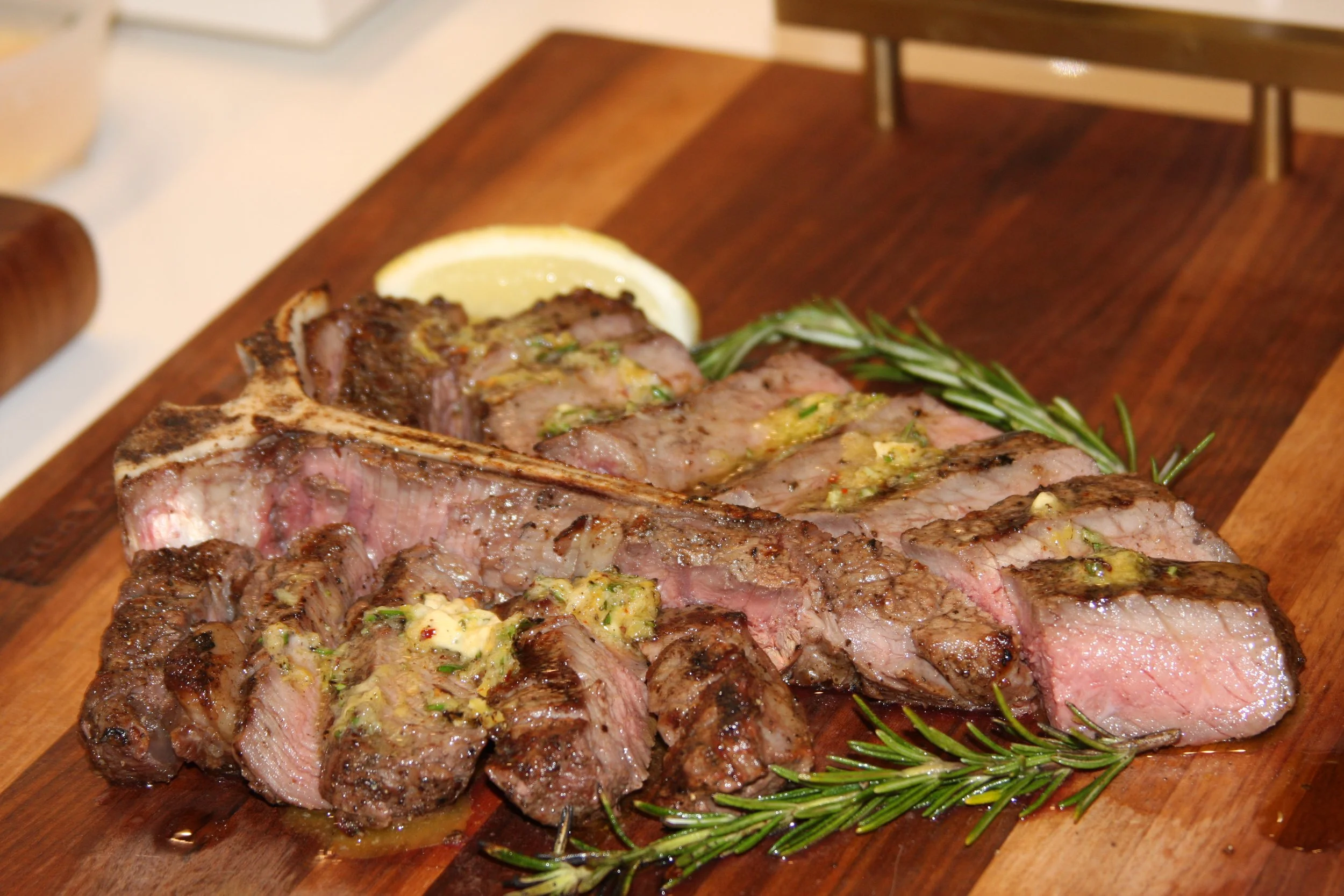 Steak cooked medium-rare with grilled seasoned garlic butter, lemon wedge, and rosemary sprigs on a wooden cutting board.