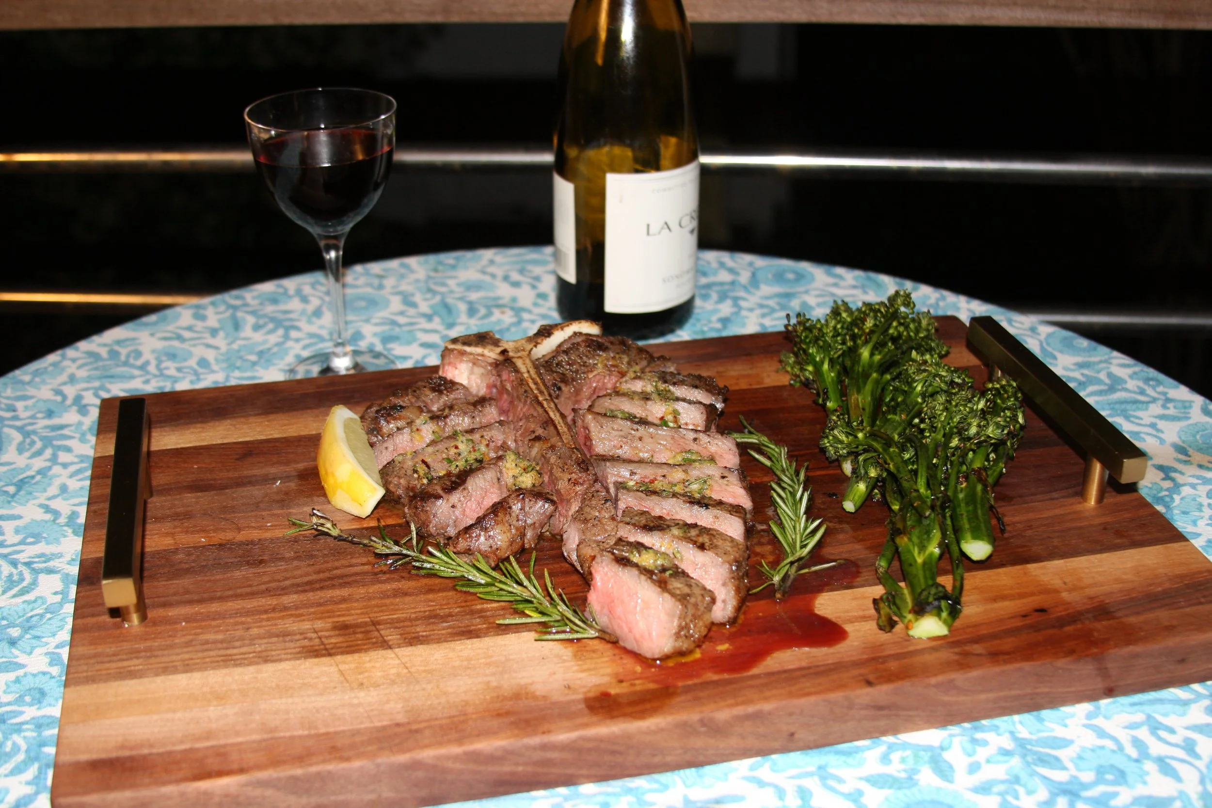 A wooden cutting board with cooked steak, a lemon wedge, fresh rosemary, and grilled broccolini, with a glass of red wine, a bottle of white wine, and a blue and white patterned tablecloth.